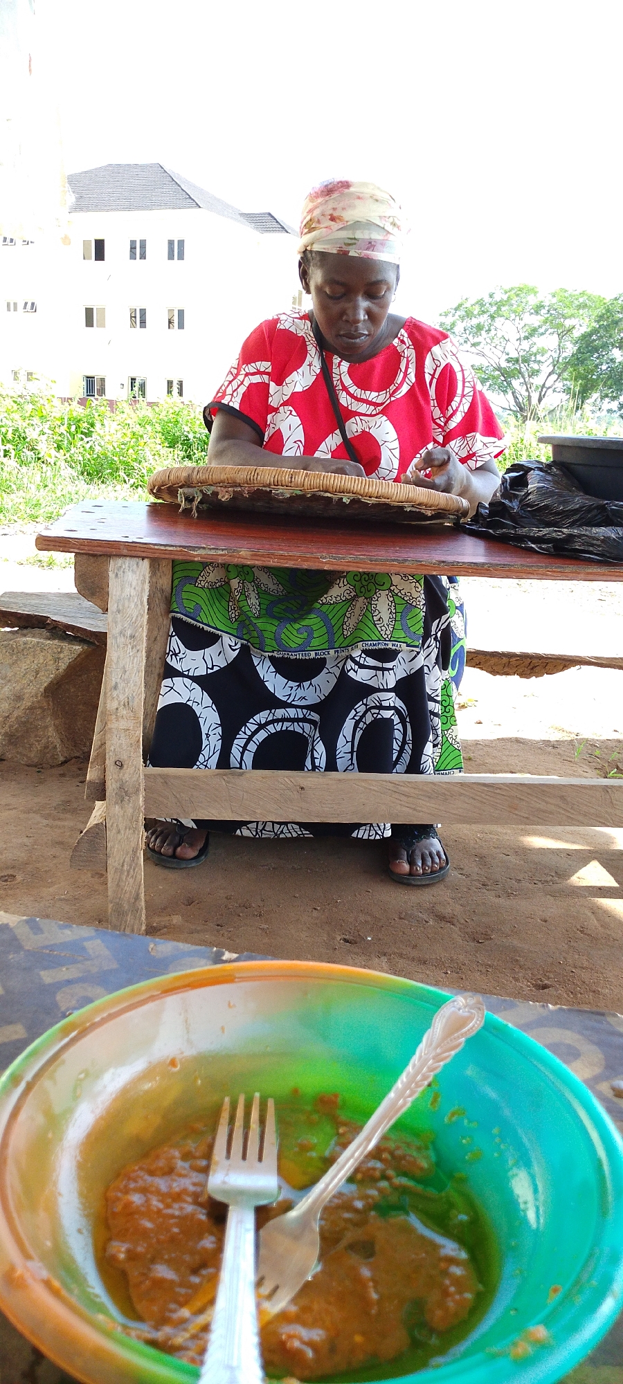 In Gumel, Nigeria, a woman wearing a red and white patterned top and a green, white, and black patterned skirt is sitting at a wooden table. She has a scarf wrapped around her head and is looking down at a woven tray in front of her, which appears to contain food. Her hands are resting on the tray, suggesting she may be preparing or serving the food. Beside the tray, on the table, is a black plastic bag. In the foreground, a colorful bowl contains a brown, saucy dish with two forks. The setting appears to be outdoors, with sandy ground and vegetation visible in the background. A white building with multiple windows is also visible in the distance.