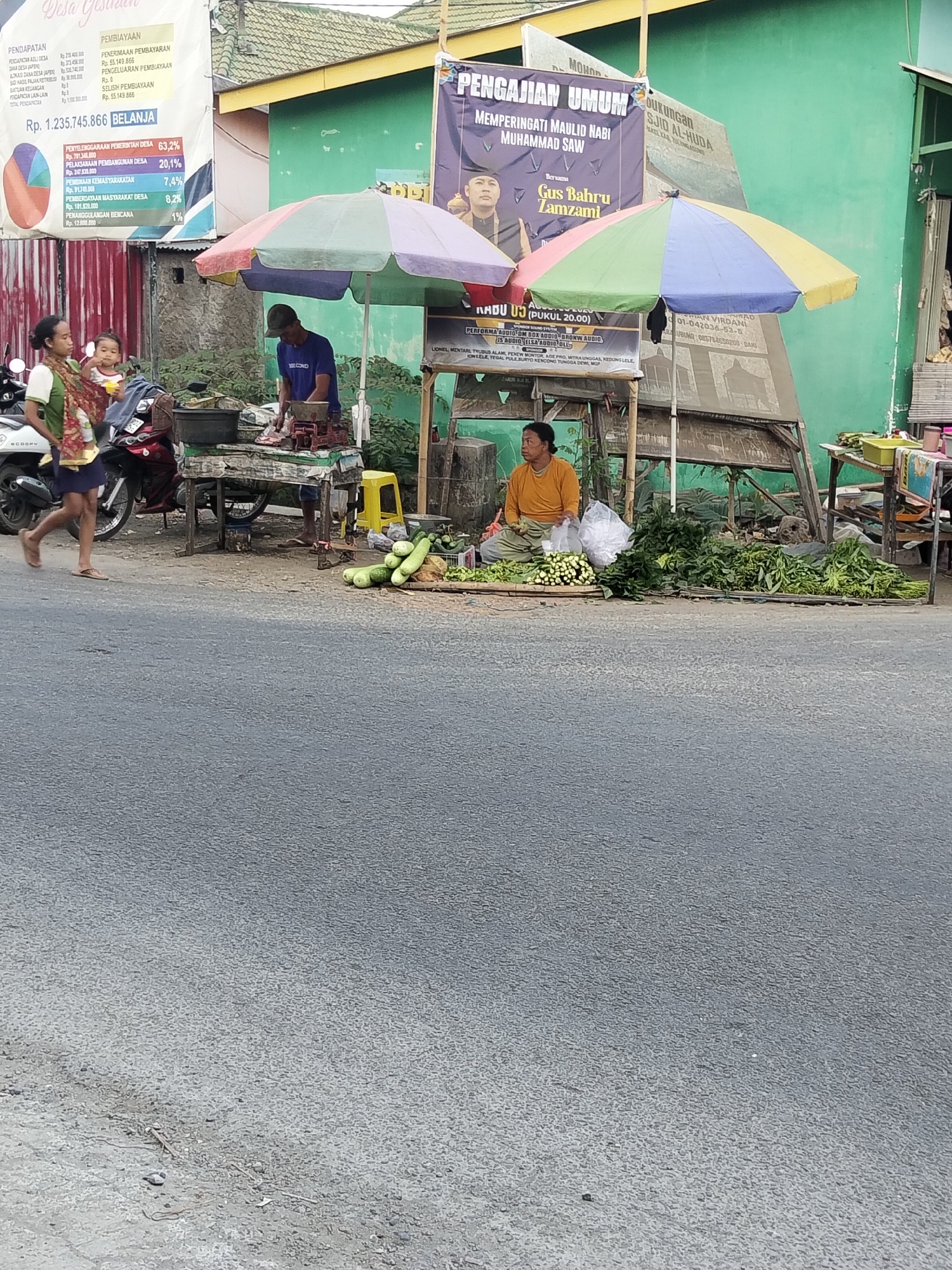 In Gempolan, Indonesia, a street scene unfolds with a vendor selling produce under colorful umbrellas. A woman and a child are walking, and a man is preparing meat at a stall. The background features buildings and posters. The image captures a snapshot of daily life in this Indonesian city.