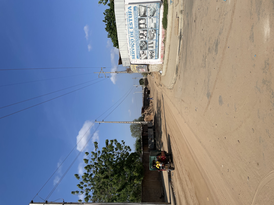 The image displays a sunny daytime scene on an unpaved dirt road in Luanda, Angola. The sky is a vibrant blue with a few wispy white clouds. Power lines and utility poles run diagonally across the frame, extending into the distance.
Along the left side of the road, a building with a white facade features a large sign. Further down, piles of dirt or debris are visible, and trees line the horizon. A green motor-tricycle, resembling a tuk-tuk, is positioned on the left side of the road, with at least one person wearing a yellow garment visible inside it. Another small, indistinct figure appears to be walking further down the road. The road surface is dry and dusty, showing visible tire tracks.
The time of day is clearly bright daylight, indicated by the intense blue sky and crisp shadows. The weather is clear and sunny.
The text visible on the building's sign, as per OCR, reads: "TAAN BI COMERCIO E SERVIÇOS (SU), LDA," followed by "SERVIÇO DE ESTUFA." Additional text includes "THA FORMACA," likely "ARTIGOS EM NOVOS," and contact numbers: "944674973-TONY / 937.912.470." The sign also displays illustrative images of vehicle interior components such as seats, steering wheels, and headrests, suggesting the business offers automotive upholstery or repair services (possibly relating to vehicle heating/cooling or interior customization, given "ESTUFA" which can mean 'furnace' or 'greenhouse' but here likely refers to vehicle interior-related service).