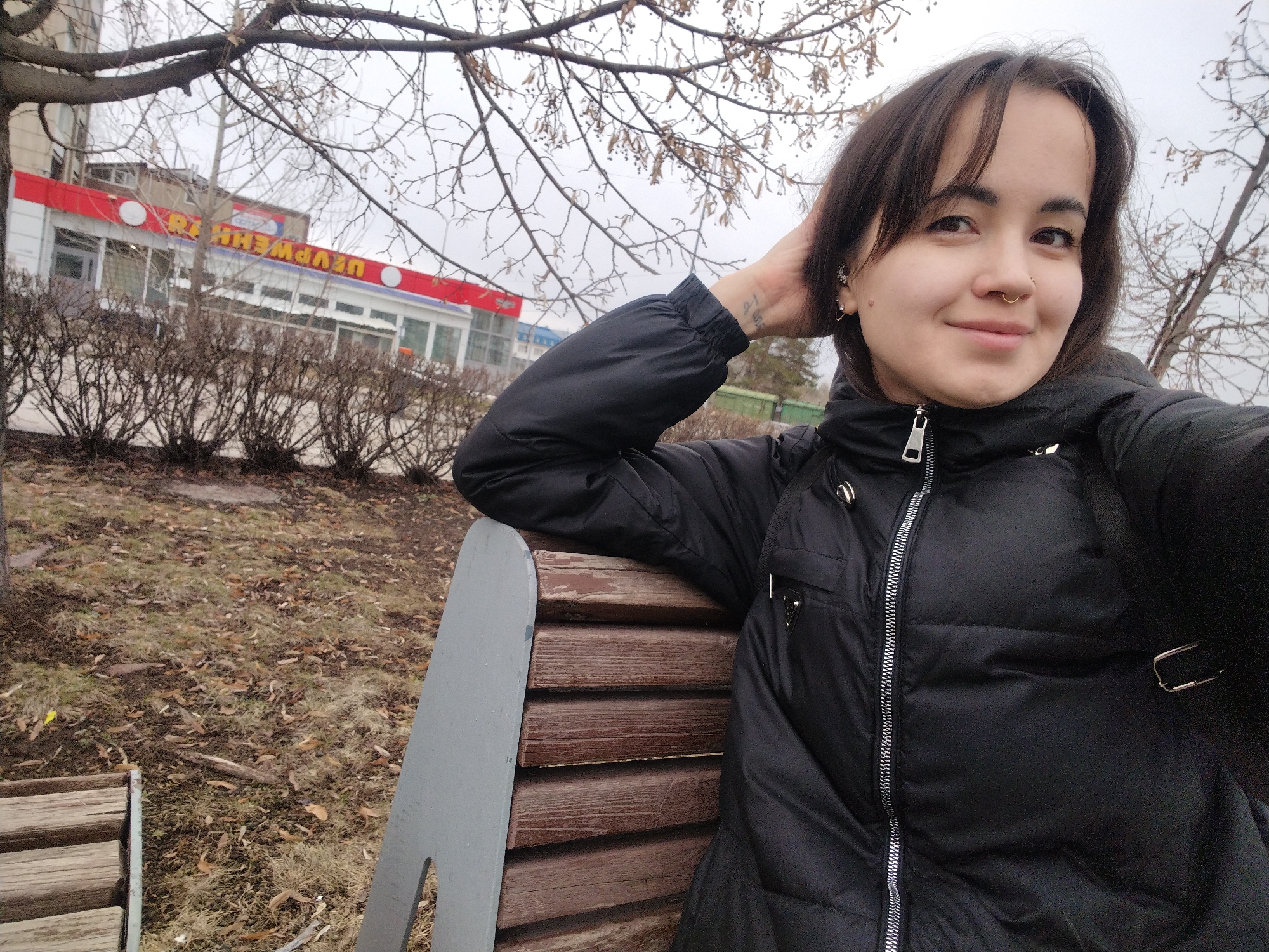 A young woman is taking a selfie while sitting on a park bench in Chaikovsky, Russia. The location is identifiable by the "Урюк-Шашлык" restaurant visible in the background. The woman has short dark hair, fair skin, a nose ring, and is wearing a black puffer jacket. Her left arm is raised, hand resting on the back of her head, revealing a small tattoo on her wrist. The park has leafless trees, bushes, and a ground covered in fallen leaves. The sky appears overcast.