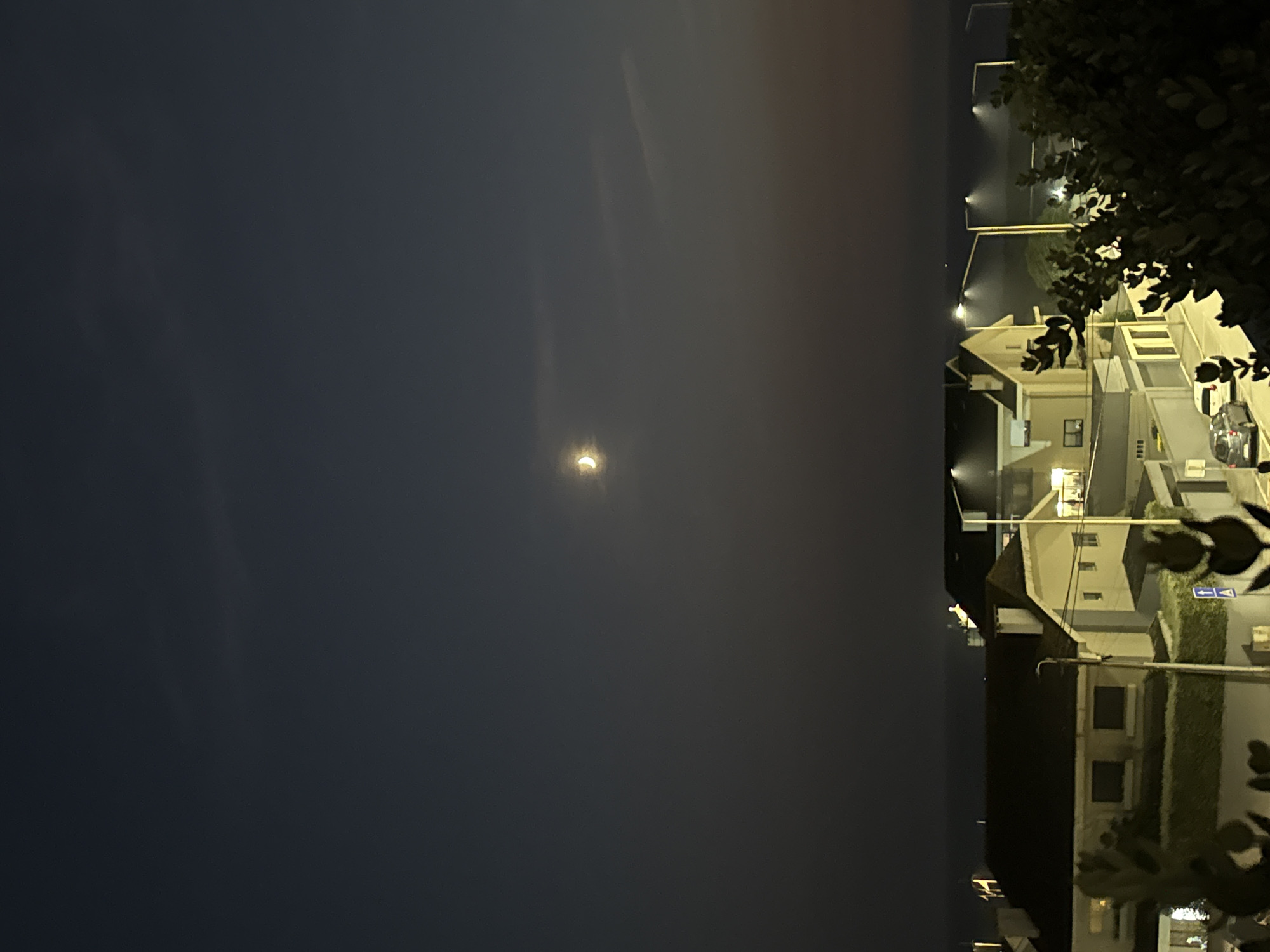 The image shows a crescent moon in a dark sky above a residential area. The sky is a deep blue fading to a lighter shade near the horizon. A few wispy clouds are visible. Below the sky, there are several buildings with illuminated windows, suggesting nighttime. The architecture appears to be of houses and possibly some apartment buildings. Streetlights are on, casting pools of light on the road and illuminating the facades of some of the structures. In the foreground, there are some dark, leafy plants, partially obscuring the view of the buildings and street. Two cars are parked on the street, one a light gray hatchback and another a white van. The overall impression is a quiet residential street at night, with the moon as a prominent celestial feature. The location is identified as Vila Nova de Gaia, Portugal.