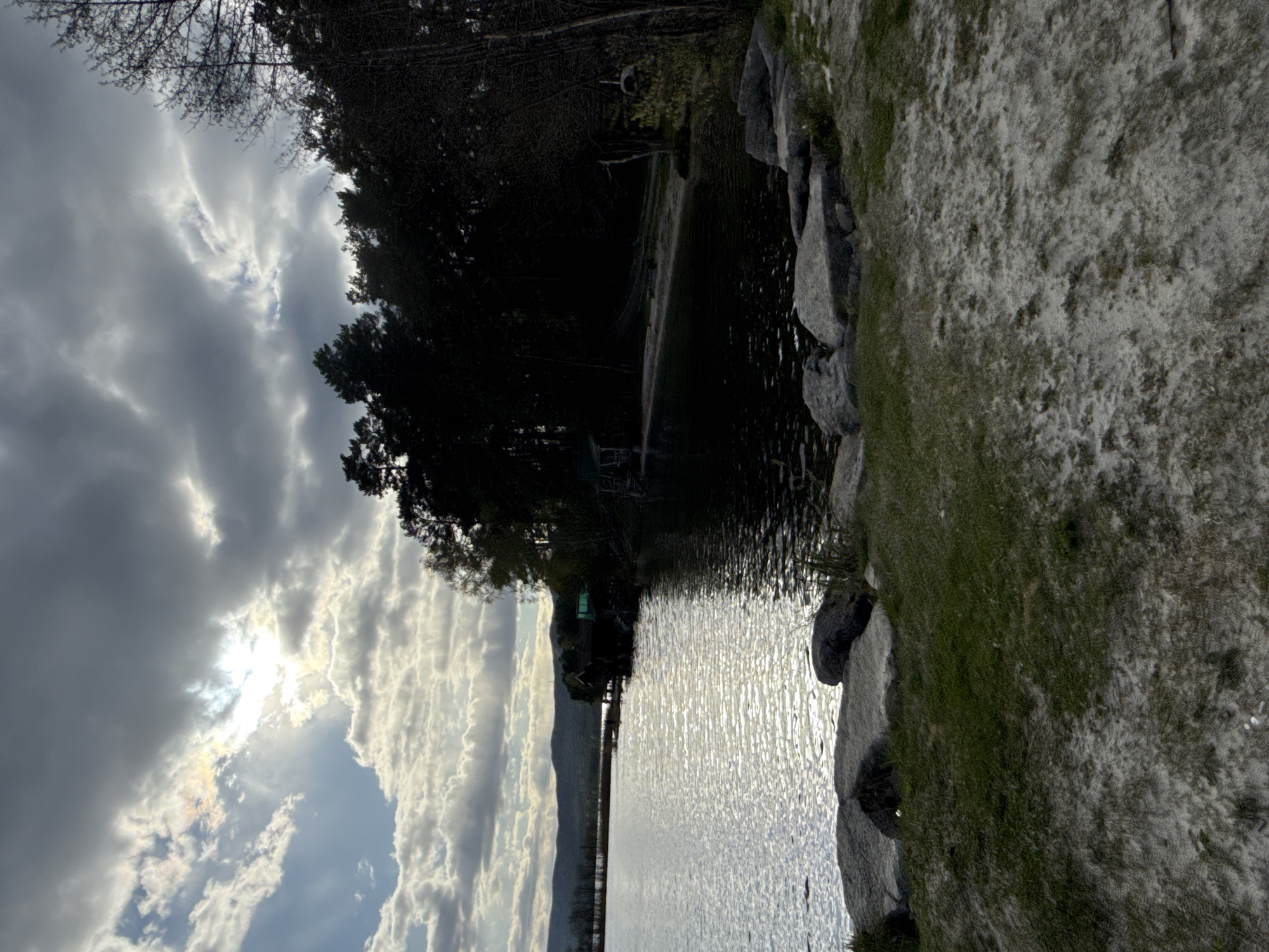 The image shows a body of water (possibly a lake or bay) with a rocky shoreline to the right. The water surface reflects the sky, which is filled with clouds obscuring the sun. Dark trees line the left edge of the water, casting shadows. No people are visible. The location appears to be a natural setting and there is no visual confirmation of its location.