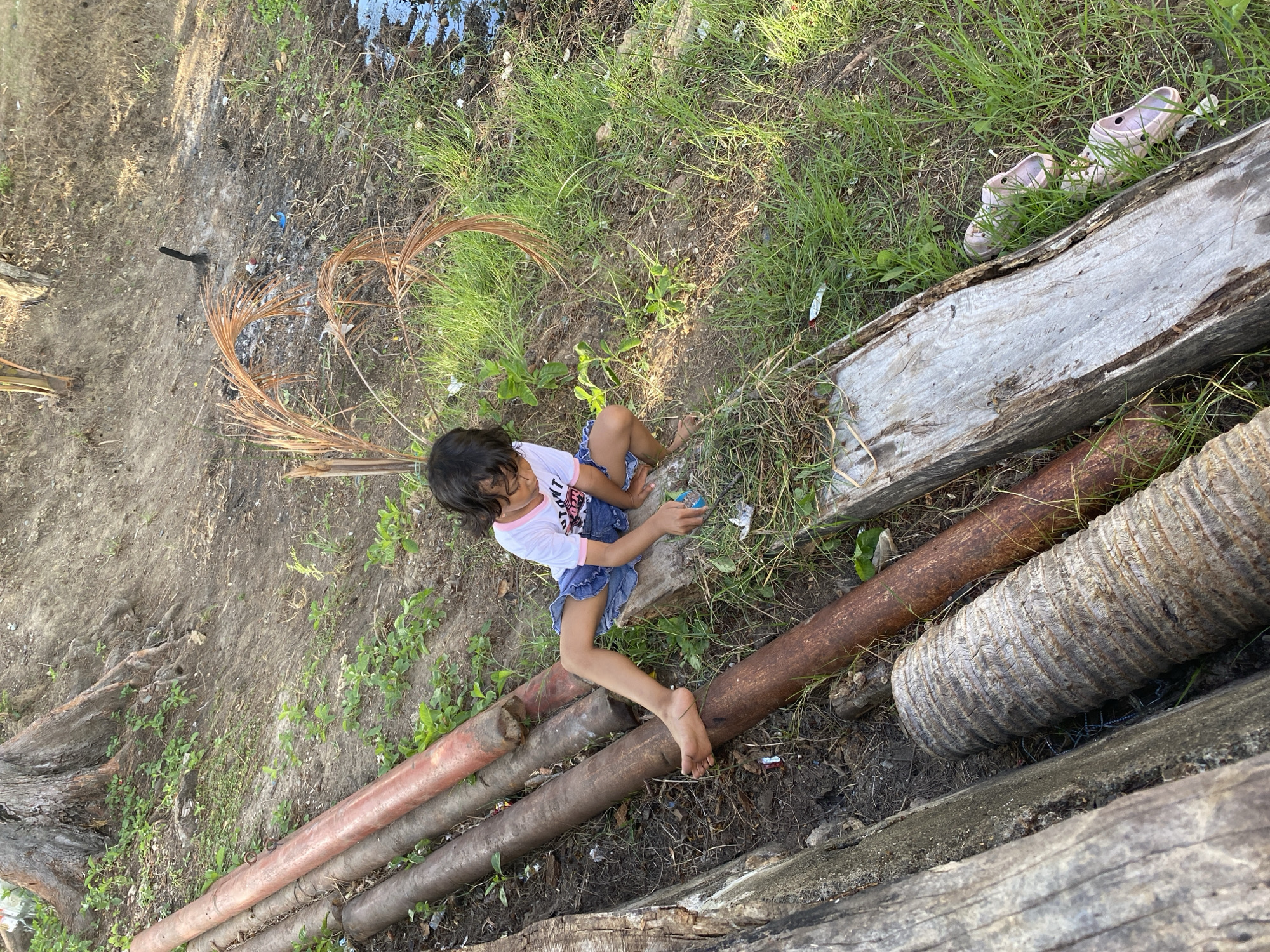 In an outdoor, possibly rural or undeveloped area, a young child with dark hair is sitting on a wooden plank, which rests on top of several large, rusty pipes. The child is wearing a white t-shirt with a graphic design and denim shorts, and appears to be playing with a small object in their hands. The child's left foot is resting on one of the pipes.
The environment is overgrown with grass and has uneven, dirt terrain. Several large, aged logs are visible, one in the foreground on the left and another diagonally in the lower right. The pipes are laid out horizontally, suggesting they might be part of an old structure or infrastructure.
In the background, a large tree stump with exposed roots is present. There is also evidence of a small fire, with ash and burnt debris scattered on the ground. A body of water, possibly a ditch or pond, is visible on the far right of the image, partially obscured by grass. A pair of light pink sandals are lying on the grass near the wooden plank on the right. The scene is illuminated by natural sunlight.