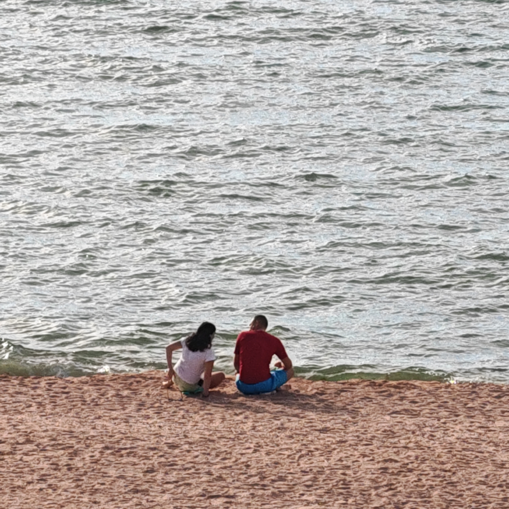 The media file depicts a wide shot of a beach with two people sitting on the sand, facing the water. The scene is set at the Мысовское сельское поселение, Ukraine, with the vast, slightly rippled water of what appears to be a sea or large lake stretching across the upper half of the frame. The beach itself is a warm, sandy color, with subtle textures indicating the granular nature of the sand.
The two individuals are positioned in the lower half of the image, sitting with their backs to the camera, both facing the water. The person on the left is wearing a white t-shirt and light-colored shorts, with dark hair. The person on the right is wearing a red t-shirt and blue shorts. Their postures suggest a relaxed, contemplative mood, possibly enjoying the view or a quiet conversation.
The water is a mix of grays and blues, reflecting the sky, and a gentle wave is visible breaking on the shore in the middle-left of the image. The lighting suggests it is daytime, with the sun likely high in the sky, casting subtle shadows. There are no visible objects other than the two people and the natural elements of the beach and water. The overall impression is one of tranquility and a peaceful moment by the water. No text is visible in the media file.