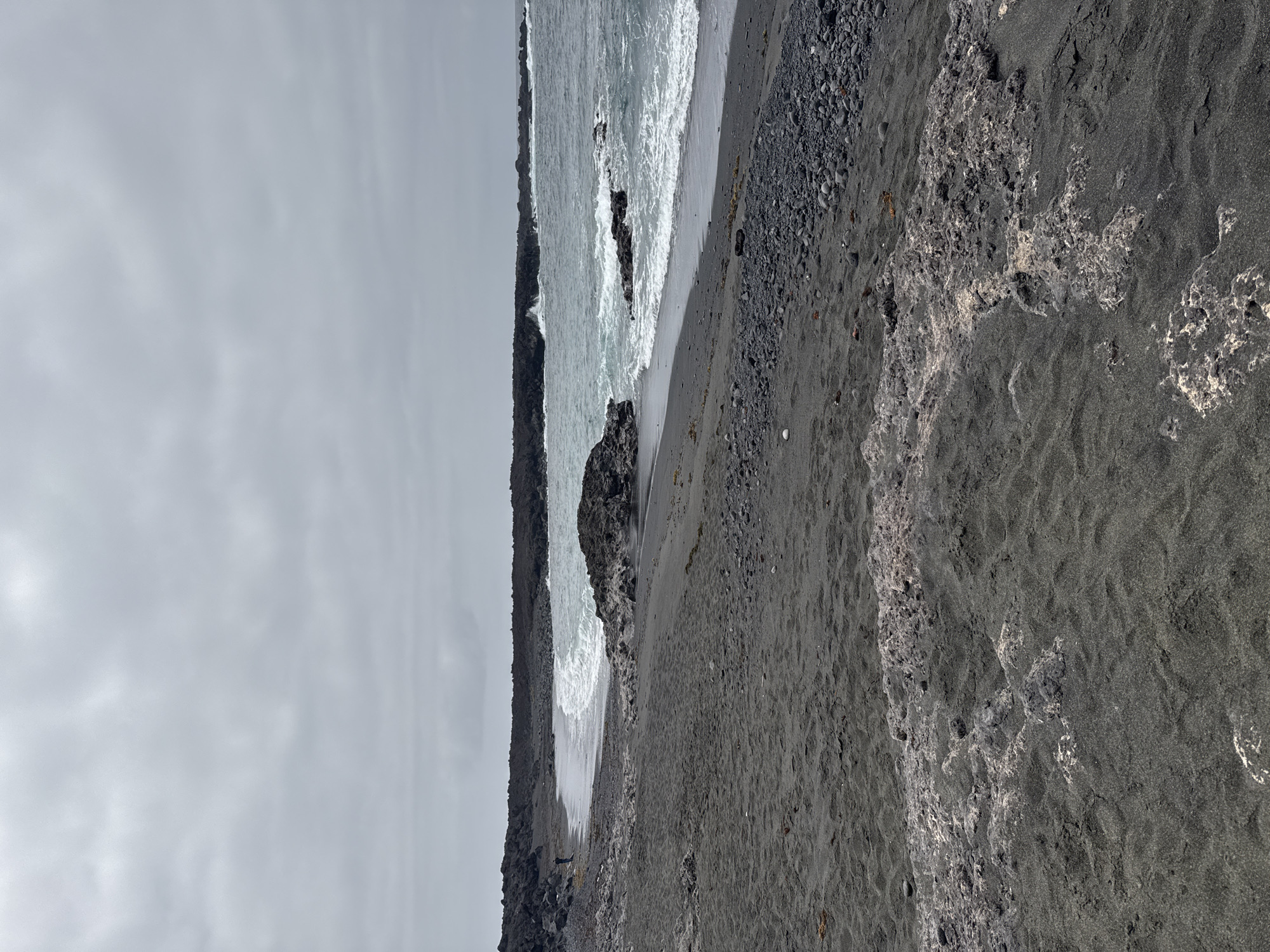 This image depicts a black sand beach in Tinajo, Spain under an overcast sky. The ocean is on the left, with waves breaking against a rocky coastline. The beach is composed of black sand and rocks, with variations in texture and patterns. There are no people visible in the image. The overall scene is monochrome due to the lighting conditions and the predominantly dark tones of the sand and rocks.