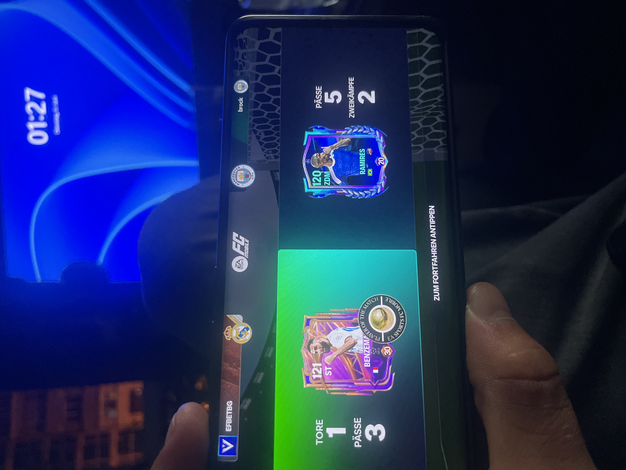 A hand holding a smartphone displays the mobile game "EA FC Mobile". The game screen shows two player cards. The left card features Karim Benzema, with stats for "Tore" (Goals) as 1 and "Pässe" (Passes) as 3. The right card shows "Ramires", with stats for "Pässe" (Passes) as 5 and "Zweikämpfe" (Duels) as 2. The game is in German, indicated by the text. In the background, a digital clock on a screen shows "01:27". The overall environment appears to be indoors with low lighting, with a hint of apartment buildings visible on the left side, suggesting an urban setting.