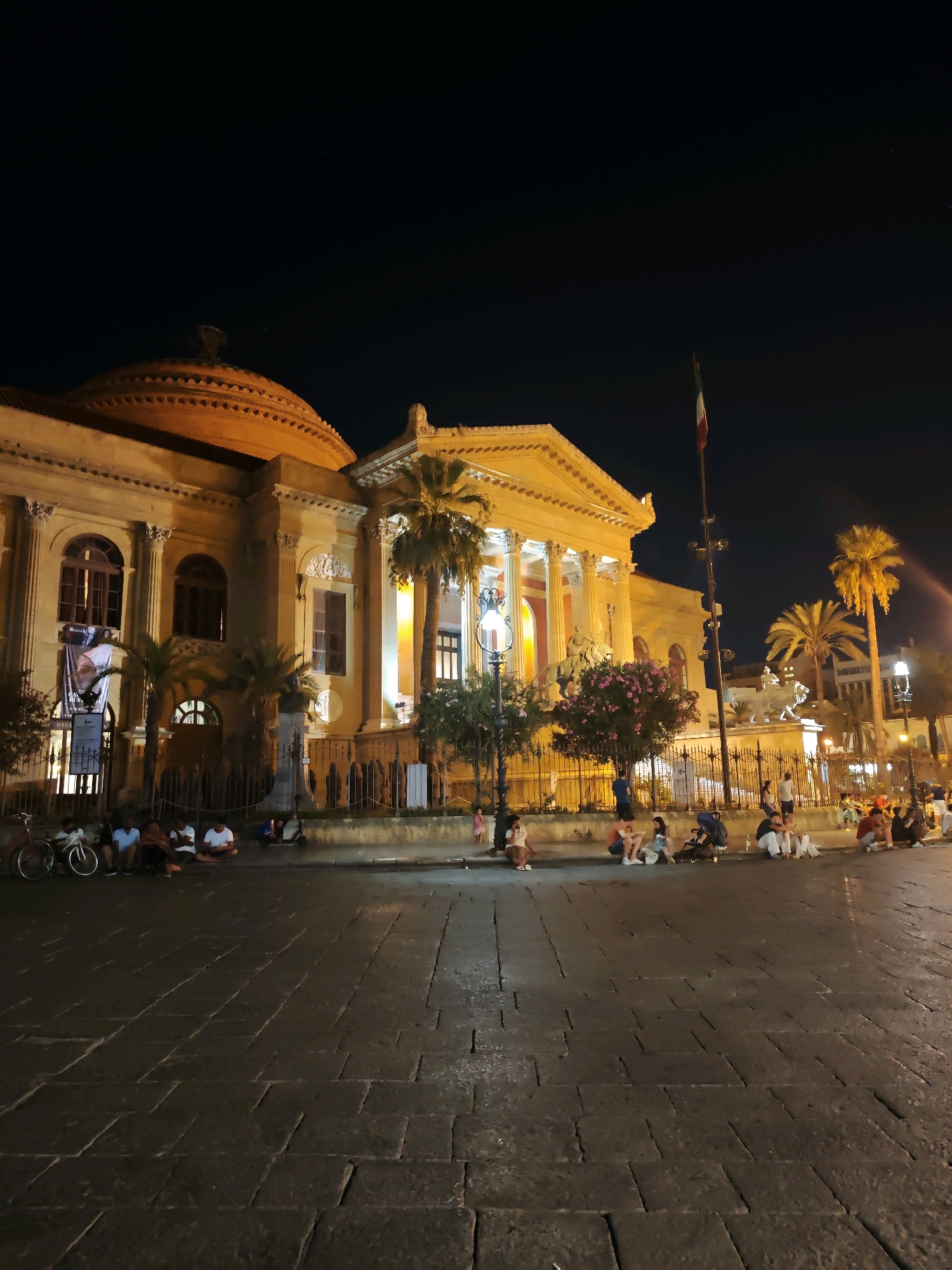 The image captures a night scene in front of the Teatro Massimo in Palermo, Italy. A group of people are seated on the stone pavement in front of the illuminated building. The theater, a grand structure with a dome and columns, is bathed in warm yellow light, highlighting its architectural details. Palm trees are scattered around the square, and a flagpole with the Italian flag stands tall. The ground is paved with large, irregular stone blocks, reflecting the ambient light. Some bicycles and a scooter are parked near the seated individuals. The sky is dark, indicating nighttime.