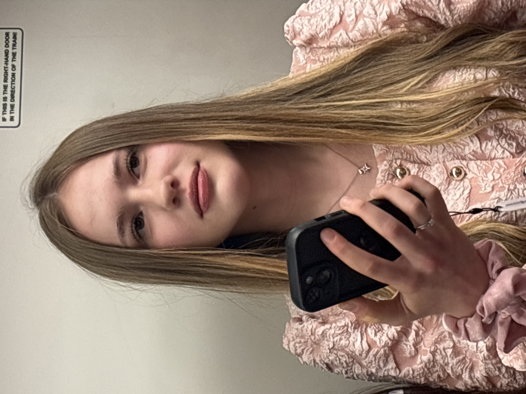 A young woman, appearing to be in her late teens or early twenties, is depicted from the chest up, positioned slightly to the left of the frame's center. She has long, straight, light brown hair with lighter highlights, parted on the side and flowing over her shoulders. Her gaze is directed slightly upwards and to the left, with a neutral expression. She wears a light pink blouse or jacket featuring a textured, possibly floral or abstract, raised pattern and metallic gold buttons down the front. A delicate silver-toned necklace with a small, star-like or geometric pendant hangs around her neck.
In her right hand, she holds a dark-colored smartphone, likely an iPhone given the visible camera array on the rear. Her thumb is positioned on the screen, suggesting active use. A silver-toned ring is visible on her right ring finger. A pink velvet scrunchie is worn on her right wrist, slightly below the hand holding the phone.
The background is a plain, light-colored wall. In the upper right corner, a white rectangular sign with black text reads: "IF THIS IS THE RIGHT-HAND DOOR IN THE DIRECTION OF THE TRAIN!" This suggests the setting is within a public transport vehicle, likely a train or subway car.