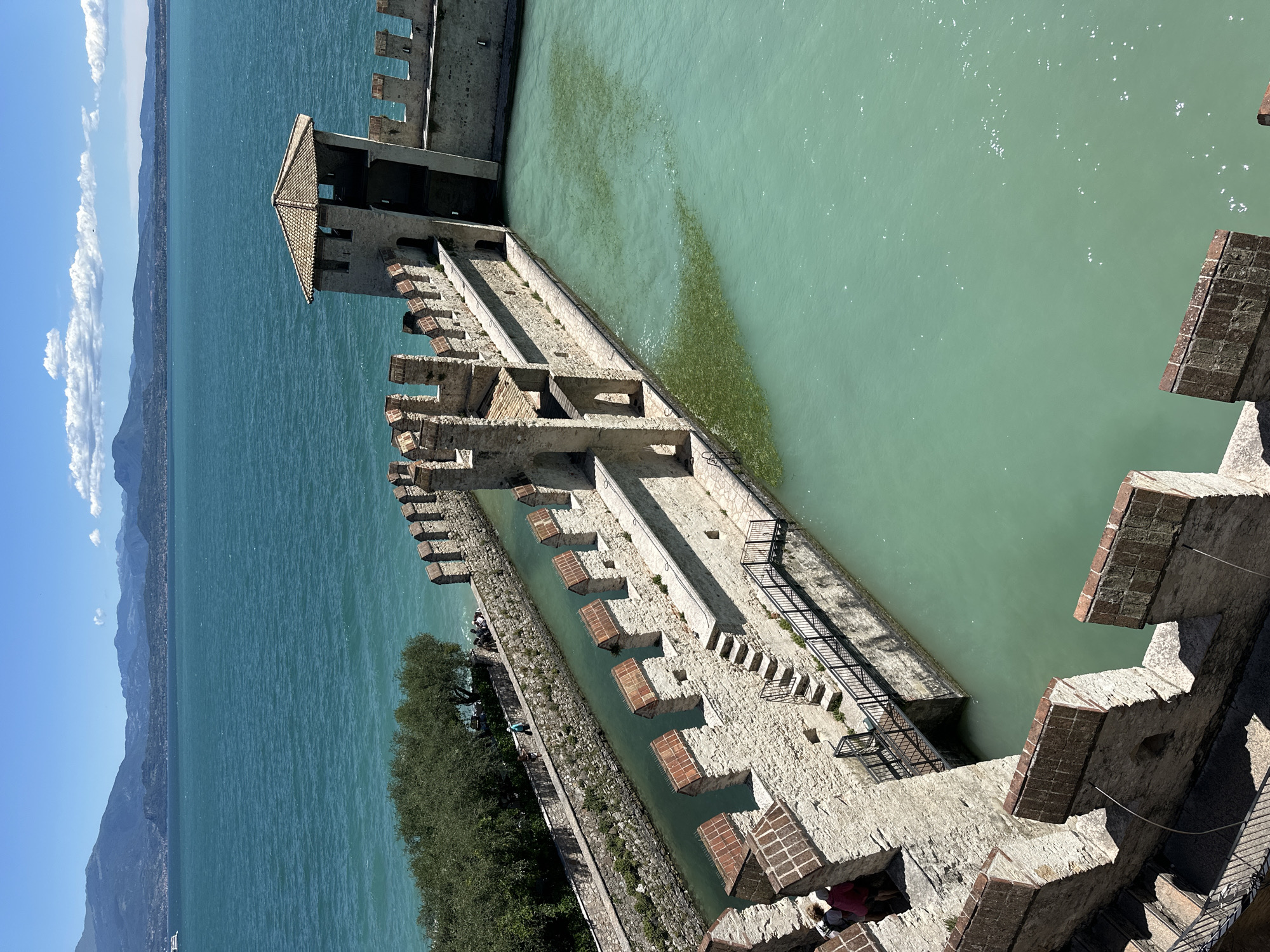 The image shows a view of the Scaligero Castle in Sirmione, Italy. The castle is a well-preserved medieval fortress located on a peninsula on the southern shore of Lake Garda. The image captures a section of the castle's stone walls, battlements, and towers overlooking a tranquil, turquoise body of water. The water appears to be a moat or a small harbor, with some vegetation visible beneath the surface. In the background, distant mountains and a clear blue sky with scattered clouds suggest a pleasant day. A few people are visible along the walkway atop the outer wall and near the entrance to the castle, suggesting it is open to visitors. The architecture is characterized by its rough-hewn stone construction and crenellated parapets.