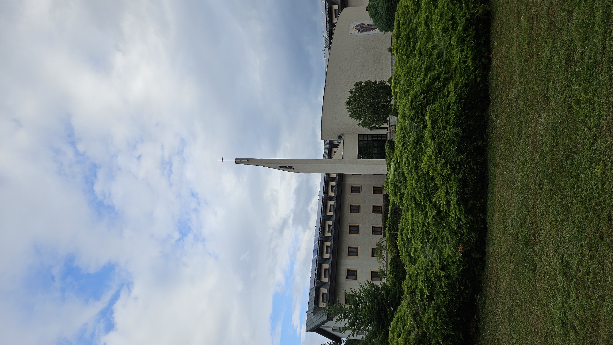 The image shows a modern building with a tall, slender tower topped with a cross. The building appears to be a church or religious institution, with a long, multi-story structure connected to the tower. Solar panels are visible on the roof of the main building. In front of the building, there is a dense hedge of green bushes and a well-maintained lawn. A small evergreen tree stands to the left of the building. The sky is partly cloudy with patches of blue. The overall impression is one of a serene and well-kept religious complex, likely located in a suburban or rural area. The provided crops do not offer any additional context regarding the specific location within Prešov, Slovakia.
