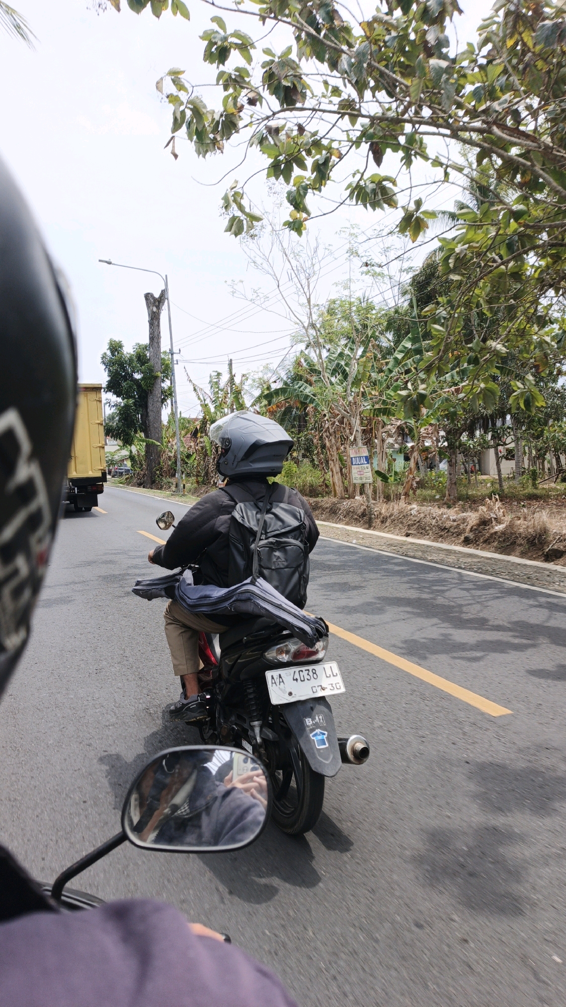 The image depicts a person riding a motorcycle from a first-person perspective, looking ahead at another motorcycle on a paved road. The rider of the leading motorcycle is wearing a black jacket, khaki pants, and a gray helmet. They have a black backpack and a long, gray bag secured to the back of the motorcycle. The motorcycle has a license plate that reads "AA 4038 LL". The road has a yellow dividing line. On the right side of the road, there is an area with dry grass, some trees, and a sign that says "DIJUAL". On the left side of the road, a yellow truck is visible. The background features trees and a utility pole with wires. The sky is overcast. The scene is likely in Sukaresik, Indonesia, given the presence of tropical vegetation and the license plate format.