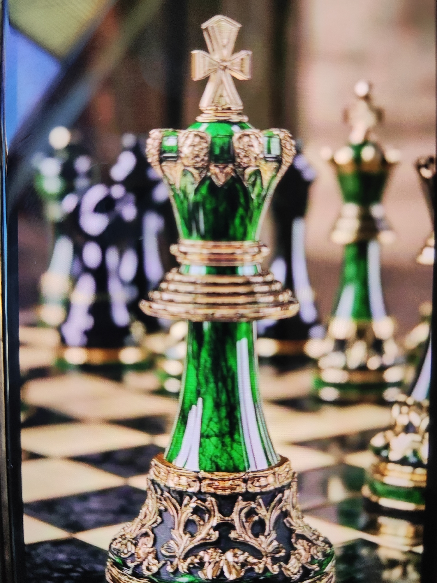 A close-up, eye-level shot captures an ornate emerald green and gold chess king piece standing prominently on a checkered chessboard. The king piece features a translucent, deep green body, meticulously adorned with intricate gold embellishments around its neck and a layered, ringed design on its mid-section. A golden cross, characteristic of a king, tops the piece. Its base is dark, possibly black, with elaborate gold filigree patterns that add to its luxurious appearance.
In the blurred background, other chess pieces of similar design, both green and gold, and darker ones (possibly black or dark grey with gold accents), are discernible. These pieces are out of focus, creating a shallow depth of field that highlights the central king. The chessboard itself displays the classic black and white (or cream) square pattern, with some squares appearing reflective.
No specific activity is underway; the scene is static, showcasing the chess set. The lighting suggests an indoor setting. There are no visible people or discernible text within the image. This highly detailed and decorative chess set, with its rich colors and metallic accents, is presented in what appears to be a sophisticated environment, set in Sochi, Russia.
