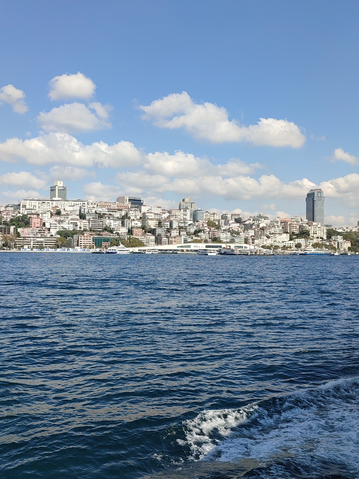 The image captures a panoramic view of the Beyoğlu district in Istanbul, Turkey, seen from the Bosphorus Strait. The foreground is dominated by the deep blue, rippling water of the strait, with a white wake trailing from the left side, indicating movement. In the mid-ground, a dense collection of buildings rises up a hillside, forming the Beyoğlu skyline. The architecture is varied, with a mix of older, white-washed structures and modern, taller buildings. One prominent tall, glass-facade skyscraper stands out on the right side of the skyline. Several boats and ferries are visible docked or moving along the waterfront. The background features a clear blue sky dotted with fluffy white clouds. The scene depicts a sunny day with a bustling urban landscape meeting the expansive waters of the Bosphorus.
