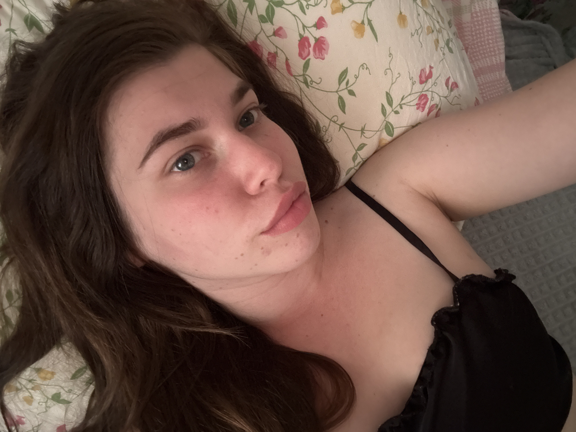 A young woman is depicted in a close-up, high-angle selfie, lying down indoors. She has long, wavy dark brown hair, fair skin, and blue eyes, looking directly at the camera with a neutral expression and slightly puckered lips. Her head rests on a white pillow adorned with a floral pattern of pink and yellow flowers and green vines. She is wearing a black bra or camisole with a ruffled detail visible on the strap and trim. Her left arm, extended towards the camera, is partially visible, indicating she is taking the photo herself. The background reveals a grey, textured blanket or bedspread and a soft pink and white checkered pattern on an adjacent pillow or throw. The lighting is soft and diffused. Minor freckles and skin imperfections are visible on her face and neck. The scene suggests a relaxed moment, likely in a bedroom in Mezhchery, Russia. No visible text is present.