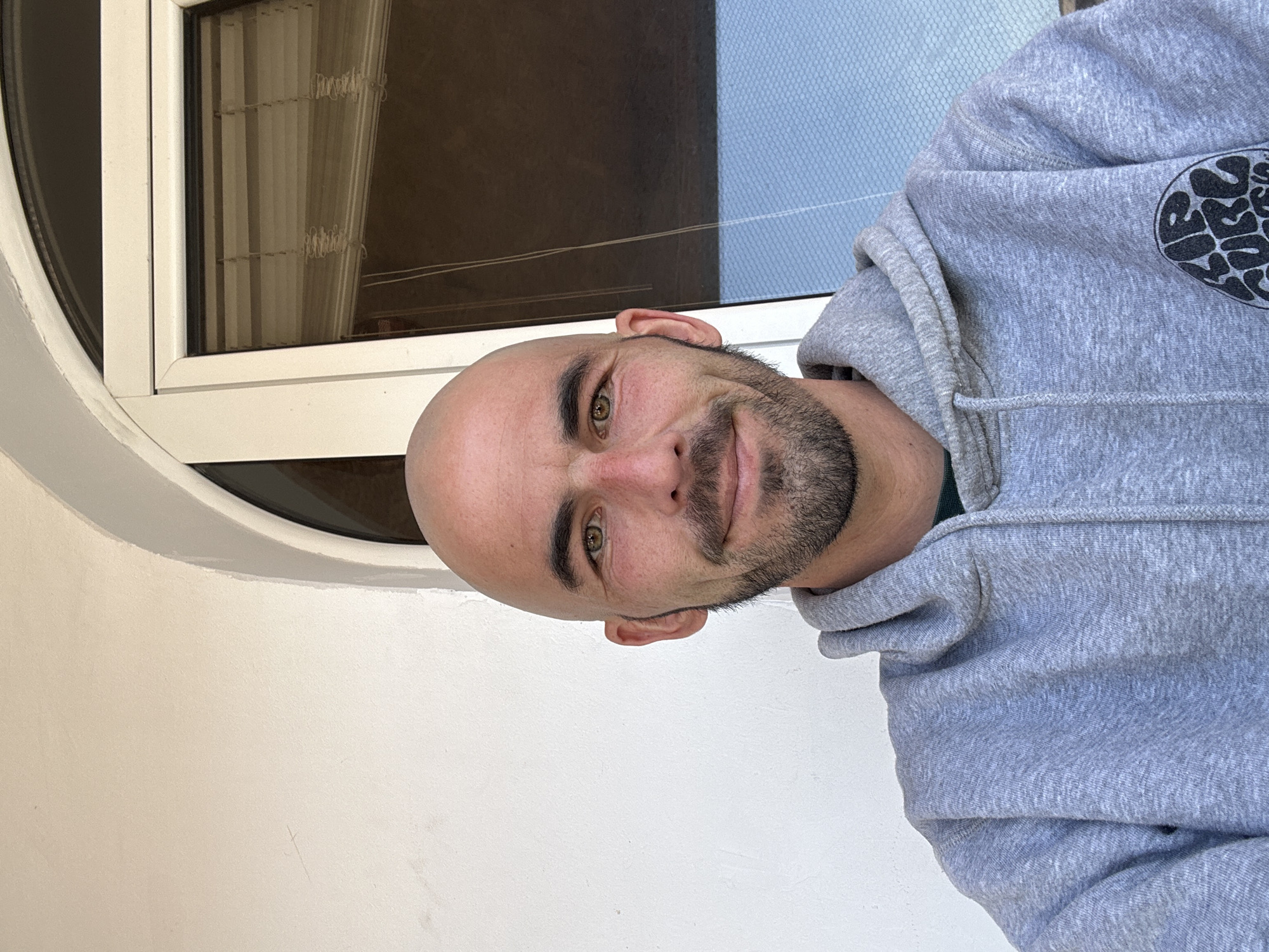 A medium shot depicts a man with a shaved head and a trimmed beard, dressed in a gray hooded sweatshirt. He is situated in front of a window in Budva, Montenegro. The window has white frames and a rounded top, and through it, there is a glimpse of both interior blinds and the exterior landscape of the city. The man is positioned against a white wall.
