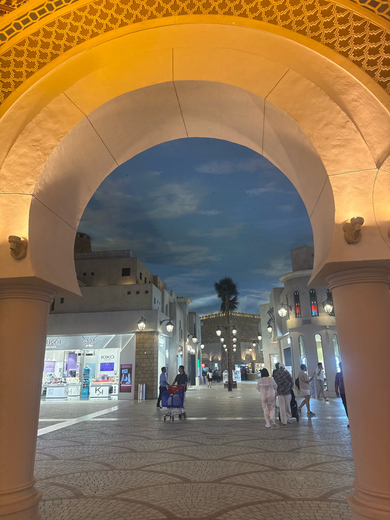 The image shows an interior view of a shopping arcade with architectural elements reminiscent of traditional Islamic design, featuring large arches, decorative motifs, and a painted ceiling that simulates a sky. The location is identified as an indoor mall in Dubai, United Arab Emirates. The scene is well-lit, likely due to artificial lighting, creating a warm ambiance.
In the foreground, a large, ornately decorated archway frames the view of the arcade. The arch's construction appears to be of stone or a similar material, painted in light, neutral tones and adorned with gold-colored geometric patterns on its upper surface. The floor is paved with patterned tiles, laid out in a sophisticated design.
Beyond the arch, the arcade stretches out, lined with shops and illuminated by street lamps that mimic traditional lampposts. The shops have modern storefronts, with one clearly identifiable as a "KIKO MILANO" cosmetics store, displaying its merchandise. The architecture of the buildings in the background draws inspiration from Middle Eastern and Mediterranean styles, with light-colored facades, arched windows, and some elements suggesting a fortified structure or tower. A single palm tree stands as a decorative element, adding a touch of exoticism.
Several people are present in the arcade, engaged in various activities. A man is seen pushing a blue shopping cart, while another man and woman walk together, appearing to be shoppers. Further down the arcade, more individuals are seen walking, some in small groups. The overall atmosphere suggests a bustling yet relaxed shopping environment. The "sky" ceiling, painted with clouds, contributes to the illusion of an open-air space within the enclosed mall.