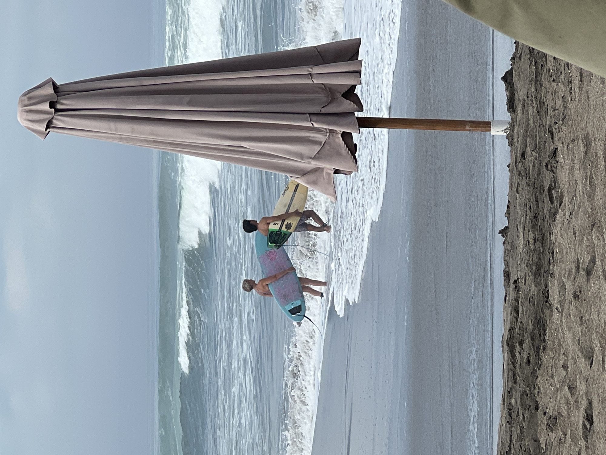 The image depicts a beach scene in North Kuta, Indonesia, with two individuals carrying surfboards walking towards the water. A large, closed beach umbrella dominates the foreground on the right side of the frame, obscuring a portion of the ocean. The ocean features moderate waves breaking on the shore under an overcast sky. The sand in the foreground appears dark and damp.