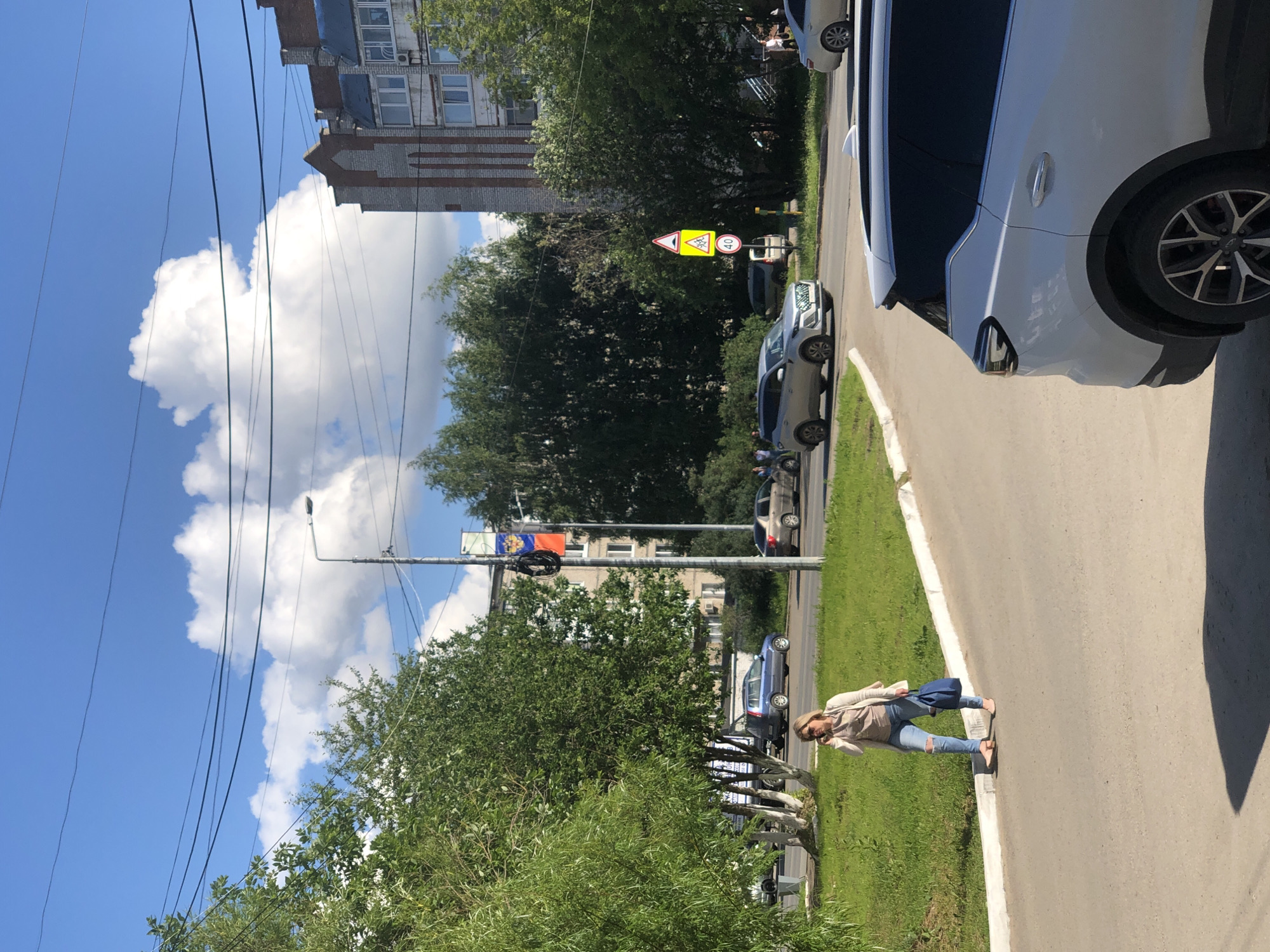 The image depicts a daytime scene on a residential street in Solikamsk, Russia. A white SUV is parked on the right side of the frame. In the foreground, a woman with blonde hair, wearing a light-colored jacket, jeans, and carrying a blue bag, walks along a grassy area next to a paved road. Several cars are visible on the road, including a silver sedan and a dark blue hatchback. Trees line the street, and a building with brick accents and apartment windows stands in the background. Overhead power lines crisscross the bright blue sky, which is dotted with white clouds. Traffic signs are visible, including one with a speed limit of 40. The overall impression is of a sunny, everyday scene in a suburban or urban environment.