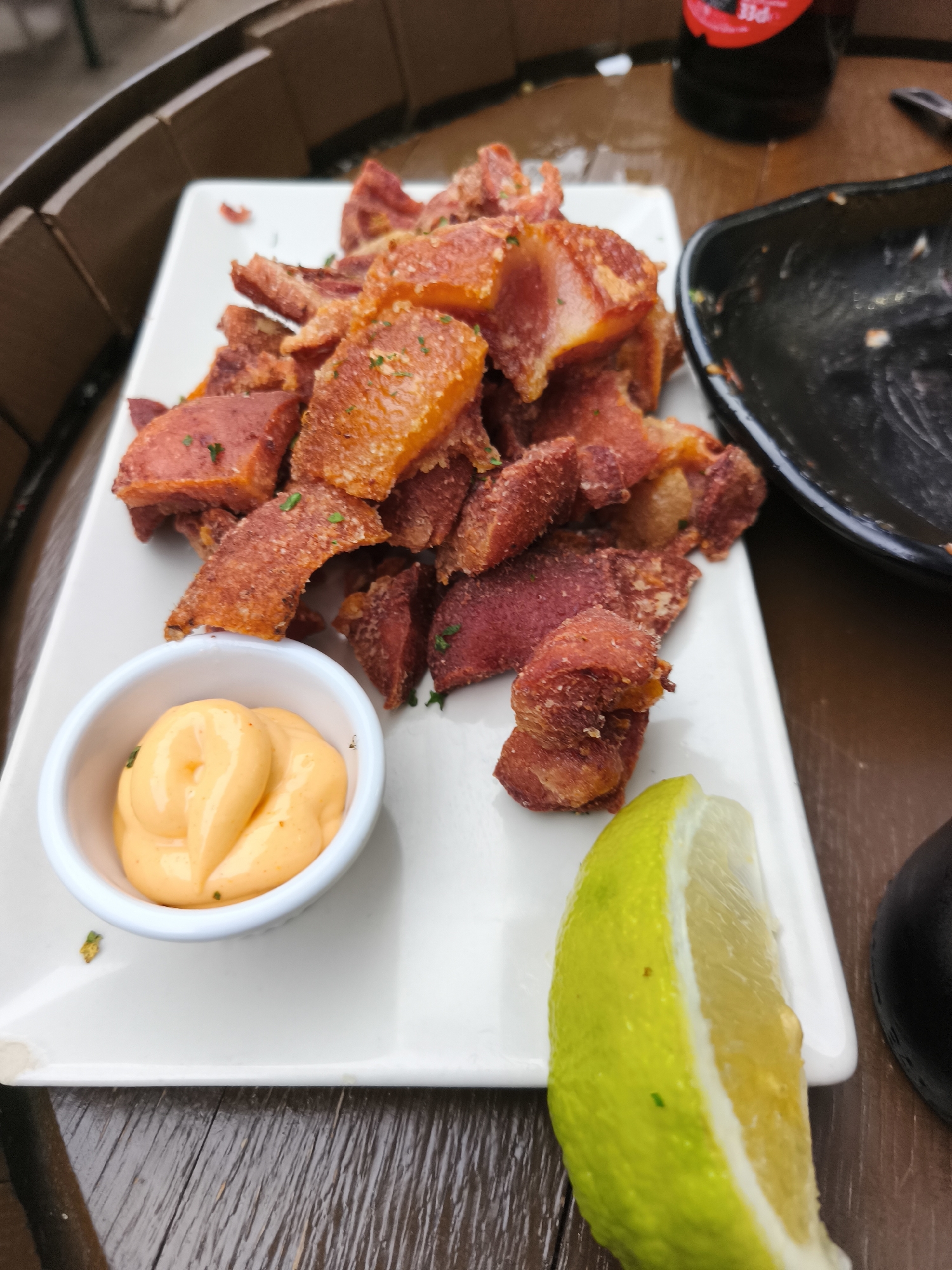 A rectangular white plate occupies the center of the image, placed on a distressed wooden table. On the plate, numerous irregularly shaped pieces of fried meat are presented, exhibiting a range of textures from crispy golden-brown to denser reddish-brown, and lightly sprinkled with chopped green herbs. A small white ceramic ramekin to the left holds a dollop of creamy, orange-hued sauce. Adjacent to the meat on the bottom right of the plate rests a fresh lime wedge, displaying its vibrant green rind and pale yellow pulp. In the blurred background, to the upper left, a curved wooden structure is visible. To the right, an empty dark-colored bowl and a dark glass bottle with a red label, partially showing "33cl" and "PIE", are discernible. The scene depicts a meal served in an outdoor or semi-outdoor restaurant setting in Picassent, Spain.