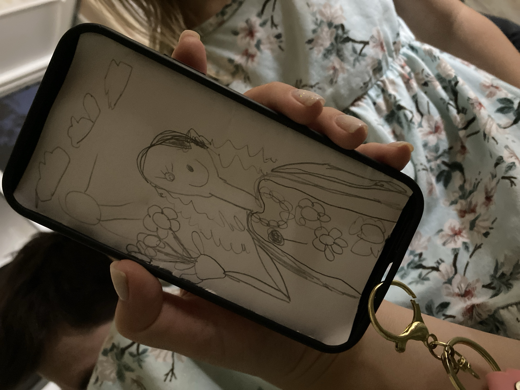 A close-up, eye-level shot shows a hand holding a smartphone with a drawing on its screen. The drawing, sketched in pencil, depicts a stick figure person with a flower bouquet and a smiling face, set against a sky with clouds. The phone is attached to a keychain with a gold clasp and a heart-shaped charm. The background features a blurred person with dark hair and a light blue floral-patterned dress. The scene is indoors, with no specific environmental context suggesting it is in Lomonosov, Russia.