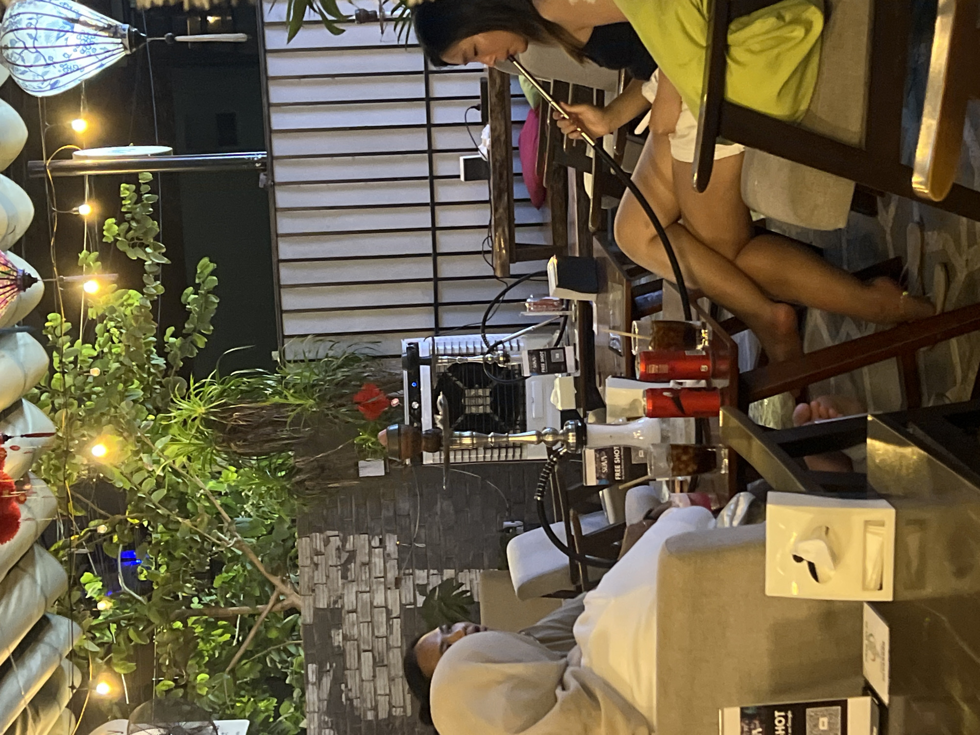 The image shows an outdoor bar or restaurant setting, likely in Đà Nẵng, Vietnam, given the presence of Vietnamese-style lanterns and architectural elements.
A woman is sitting in a chair and smoking a hookah. The hookah stands on a table next to her. Several glasses and promotional materials for alcoholic beverages are also present.
Another individual is seen reclining on a cushioned seating area nearby. There are decorative plants and string lights in the foreground, adding to the ambiance.
The setting includes wooden slats and brickwork, creating a semi-enclosed outdoor space.