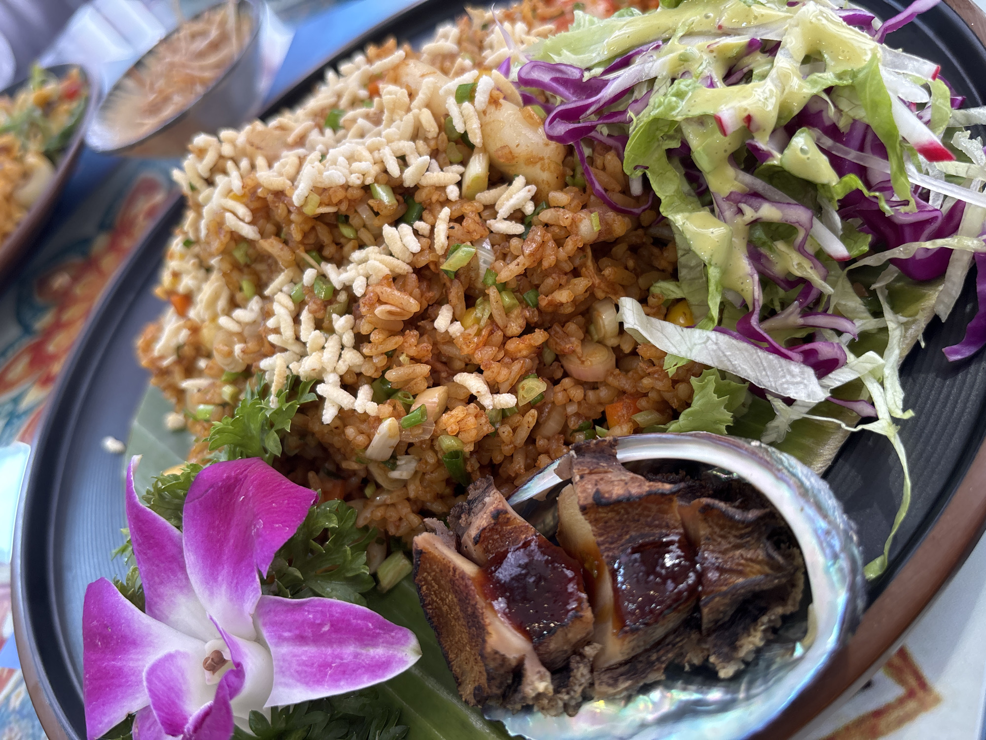 A close-up shot shows a plate of fried rice in a restaurant in Đà Nẵng, Vietnam. The fried rice is topped with white puffed rice and mixed with small pieces of vegetables and what appear to be seafood. To the right is a salad with shredded lettuce, red cabbage, and a pale green dressing. At the bottom, a portion of cooked and sliced abalone rests in its iridescent shell. An orchid flower lies near the edge of the plate. The food is served on a dark-colored plate. A small bowl containing food is partially visible in the background. The table is covered with a patterned tablecloth.