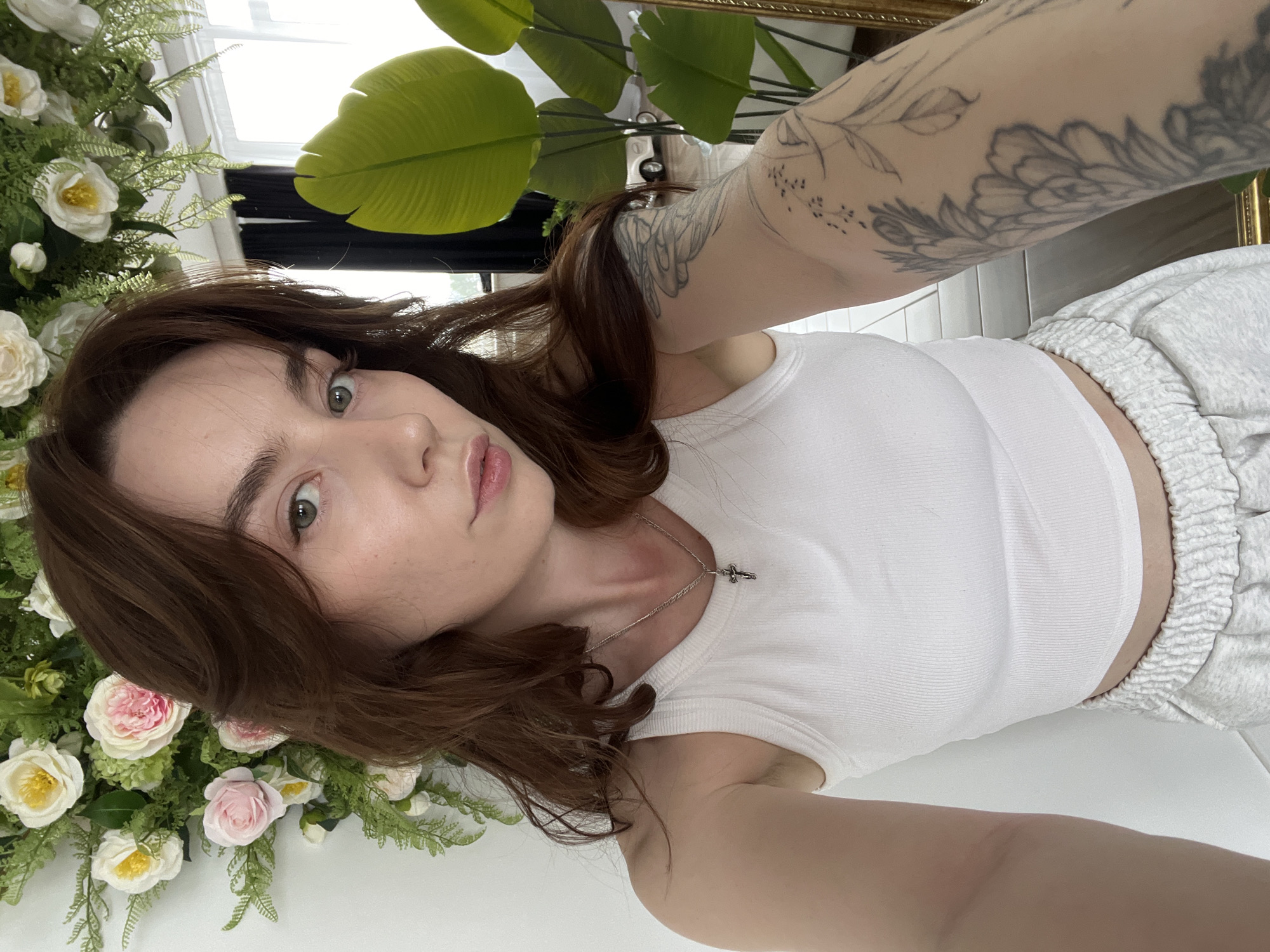 A young woman with shoulder-length auburn hair, green eyes, and fair skin takes a selfie indoors. She wears a white ribbed tank top and grey sweatpants. A silver chain with a cross pendant is visible around her neck. Her left arm, adorned with a floral tattoo, is extended toward the camera. A floral arrangement of white and pink roses is on the left side of the frame. A large, green plant is visible behind her. The location is not discernible.