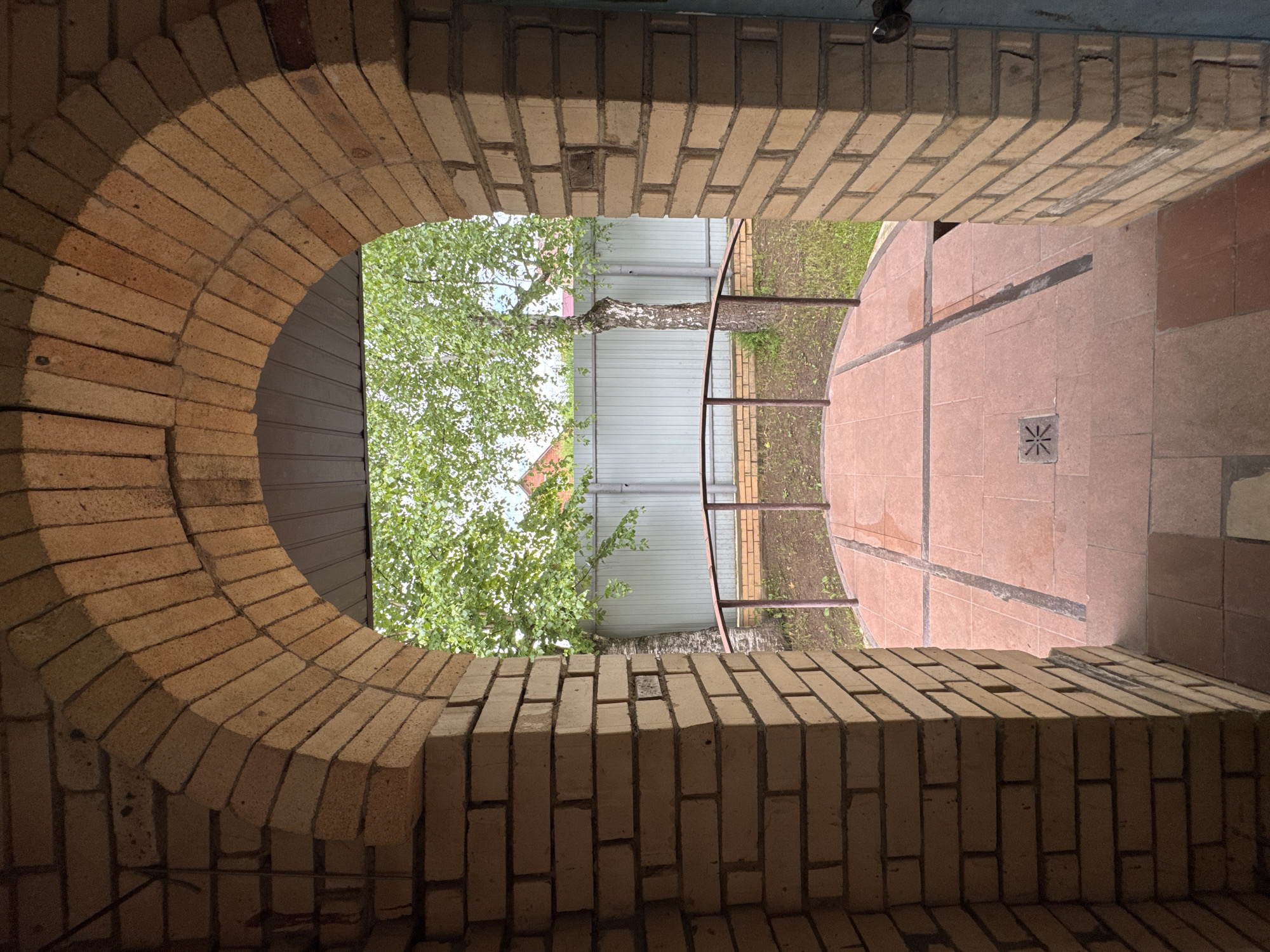 The image depicts an arched brick entryway leading to an outdoor area with a tiled patio and a metal railing. Beyond the patio, a grey fence encloses a yard with a grassy area and a tree with green foliage. The brickwork of the archway is light tan and appears weathered. The patio tiles are reddish-brown, and a drainage grate with a star pattern is visible in the center. The overall scene suggests a residential outdoor space, possibly a backyard or garden area. The location is identified as Naro-Fominsk, Russia, but no specific context or individuals are present in the image.