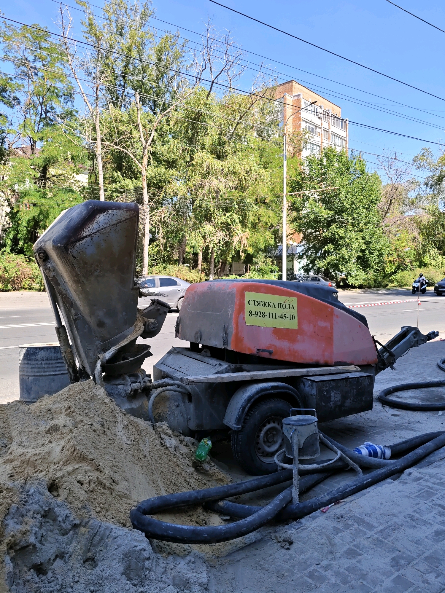 The image depicts a construction site on a sunny day in Rostov-on-Don, Russia. In the foreground, a large mechanical device, likely a concrete pump or screed machine, is parked on a sidewalk. The machine is predominantly orange and gray, with visible hoses and a pile of sand and construction debris next to it. A large metallic bucket, also part of the machinery, is angled upwards.
Behind the construction equipment, a street is visible with traffic, including a silver sedan and a motorcycle with a rider. The background is filled with lush green trees and a multi-story brick building, typical of urban architecture. Overhead power lines crisscross the blue sky. The scene suggests ongoing construction or repair work in a city environment. No people are clearly depicted in action, though the presence of a motorcycle rider implies activity. The overall impression is of a typical workday in a residential or commercial area undergoing renovation.
