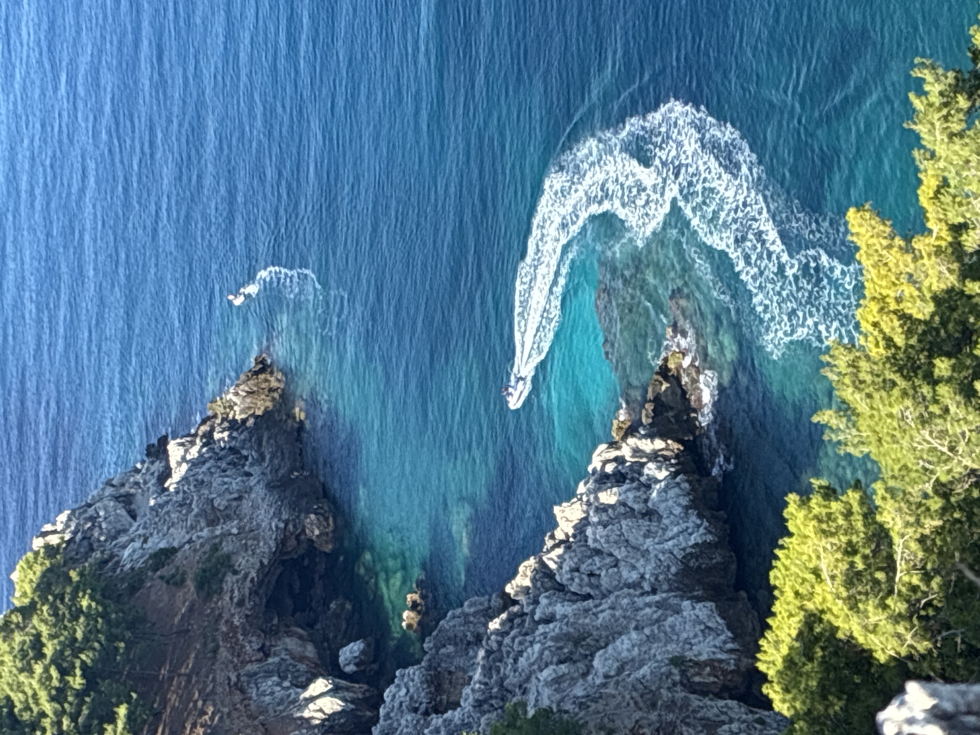 The image depicts a rugged, rocky coastline with lush green vegetation in the foreground, overlooking a vibrant turquoise sea. The water is clear enough to reveal submerged rocks near the shore. Two boats are visible in the water. One is a jet ski with a person on it, creating a white wake as it maneuvers away from the shore. Further in the distance, a smaller, white boat with a person on it is also present, leaving a faint wake. The rocky cliffs have areas of shadow, suggesting the sun is high in the sky. The scene is consistent with the coastline of Alanya, Turkey, known for its dramatic cliffs and clear Mediterranean waters.