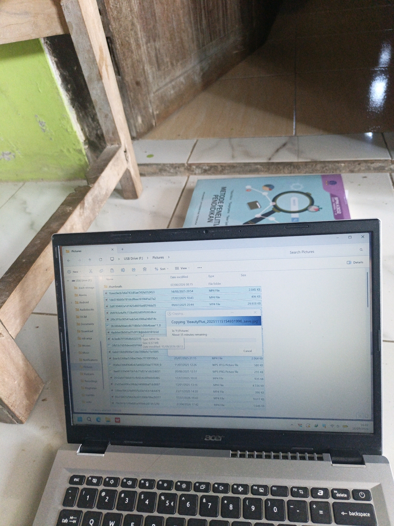 In the city of Kamulyan, Indonesia, an open laptop is positioned on a tiled floor, with a portion of a wooden chair and a wall painted green visible in the background. The laptop screen displays a file explorer window, showing a list of files and folders on a USB drive, with a file copy operation in progress titled "Copying 'BeautyPlus_20251115154931996_save.jpg'". The laptop's keyboard is visible in the foreground.