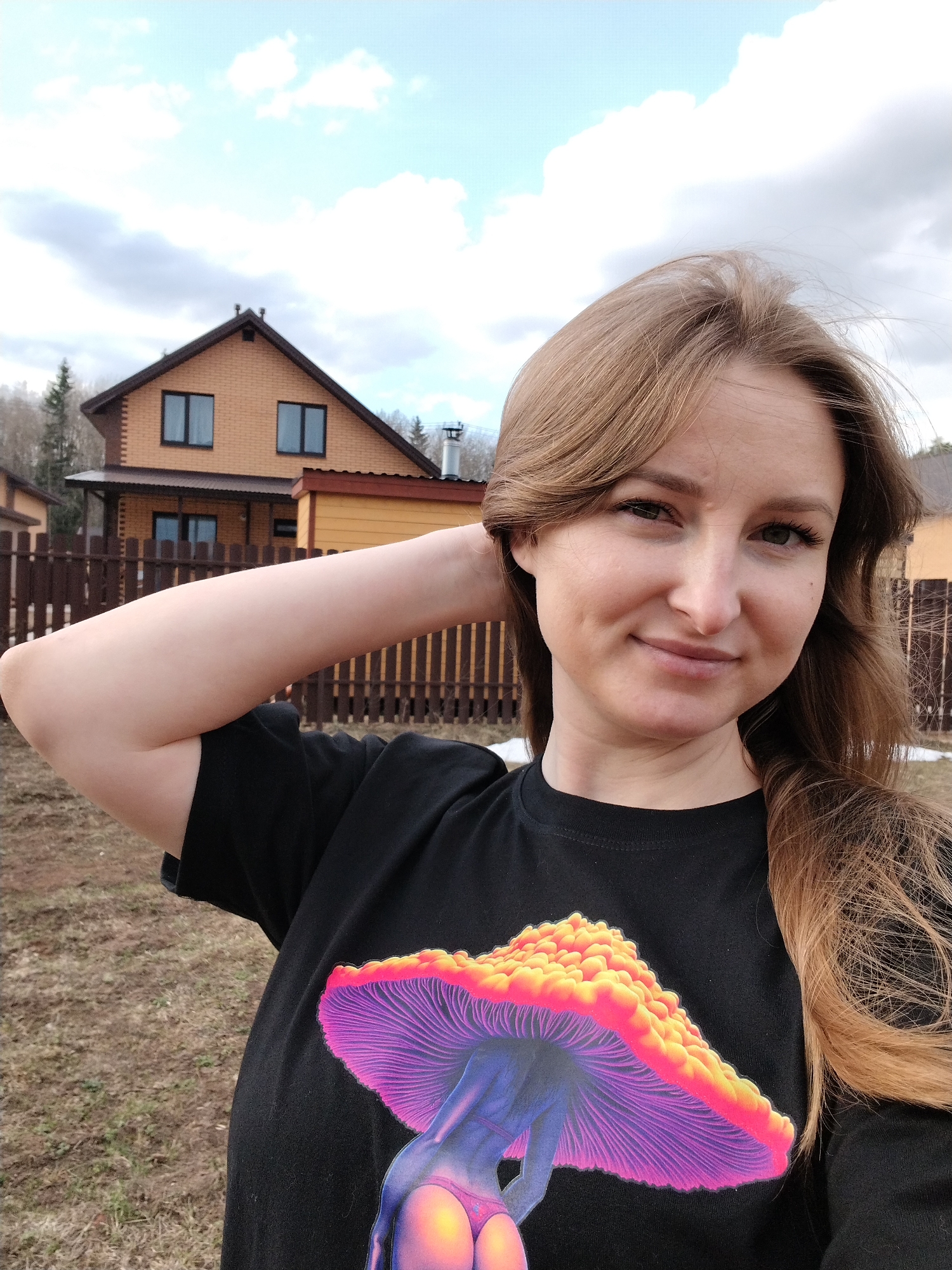 A young woman is posing in a selfie in front of a wooden fence and a house in what appears to be a suburban area. The house behind her is constructed from brown brick and has a brown roof. The sky is overcast, with gray and white clouds. The ground is covered in grass and some bare patches of dirt. The woman is wearing a black t-shirt with a brightly colored design of a mushroom on it. She has light brown hair and is smiling slightly. Her right arm is raised, with her hand touching her hair. The location is Chaikovsky, Russia.