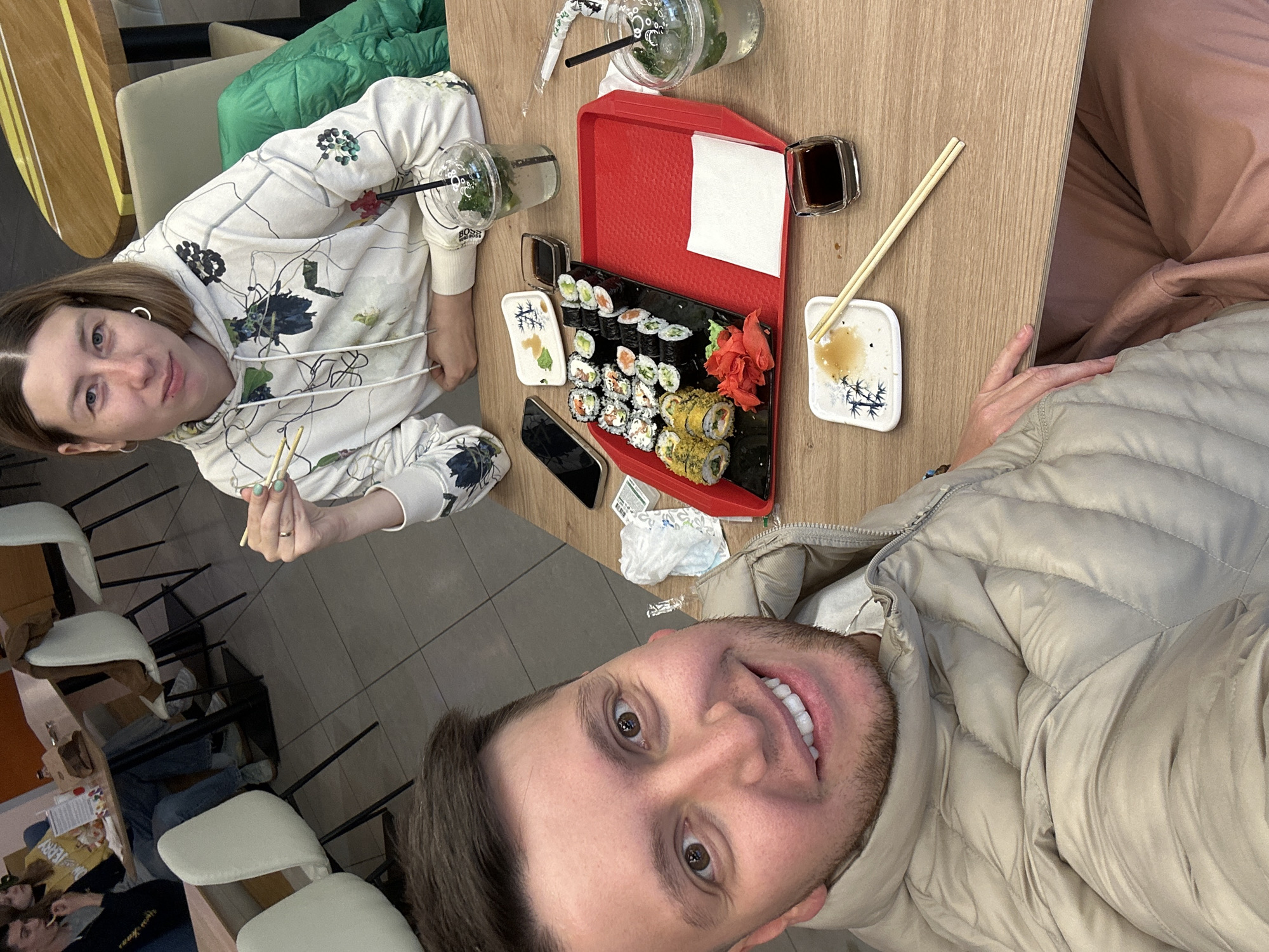 A man and woman are sitting at a table in what appears to be a restaurant. A red tray with sushi, ginger, soy sauce, and beverages is on the table. The woman is holding chopsticks. A smartphone is placed on the table next to the sushi. The man is facing the camera, taking a selfie. In the background, there are chairs and tables, suggesting a public eatery. Considering the language and architecture generally found in images online, and without specific identifying marks, it could be anywhere. Without further information, it's impossible to confirm the city as Moscow, Russia, but it is a plausible location given the attire and the type of food.