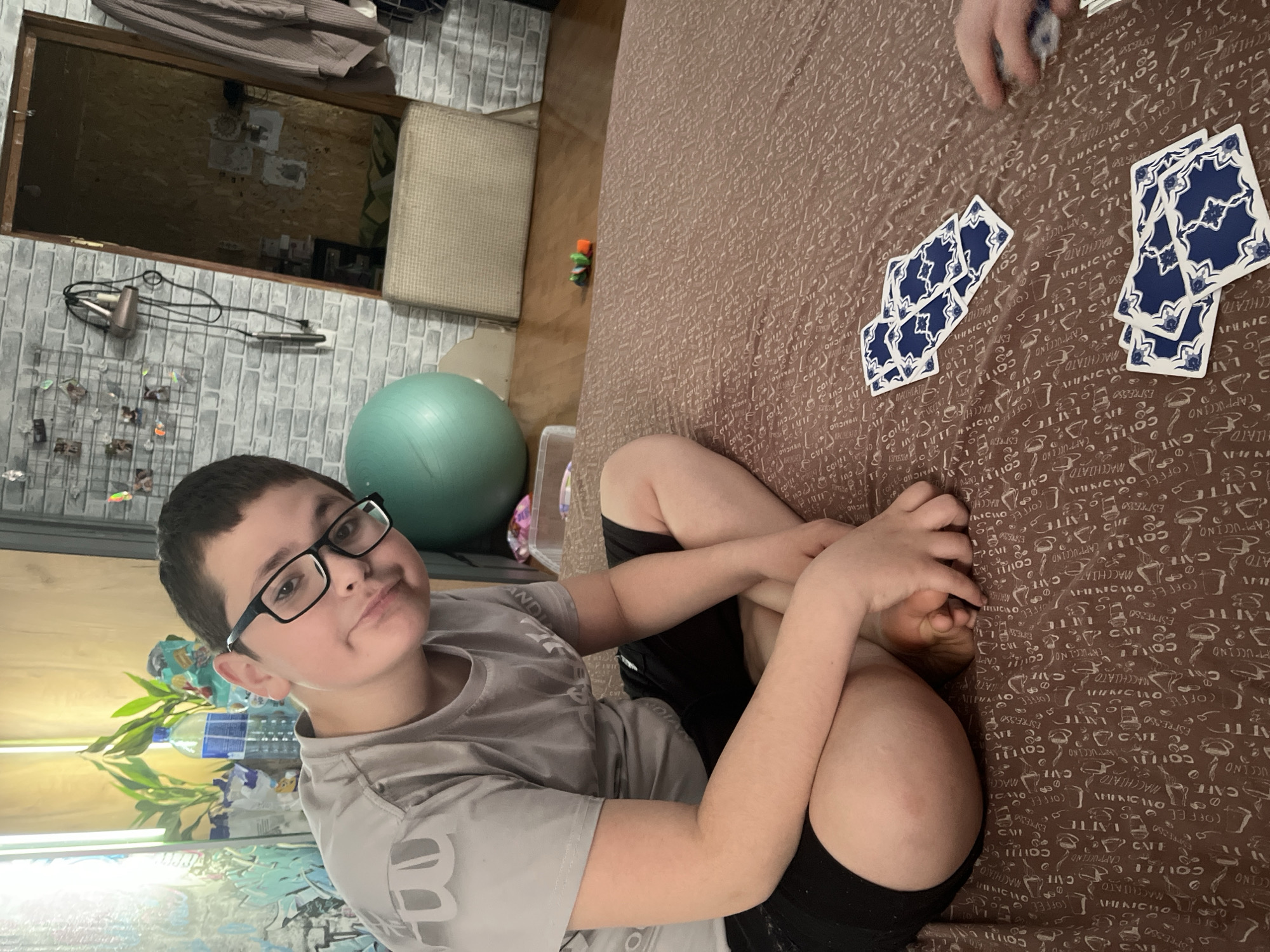 A boy with dark hair and glasses sits on a bed with a brown, patterned cover. He is wearing a gray t-shirt and black shorts. His legs are crossed, and he is holding his foot. Playing cards are spread out on the bed near his legs. The background shows a room with brick-like wallpaper, a window, a green exercise ball, and a plant. The location is likely indoors. The image does not contain sufficient information to verify its location as Vesyoloye, Russia.