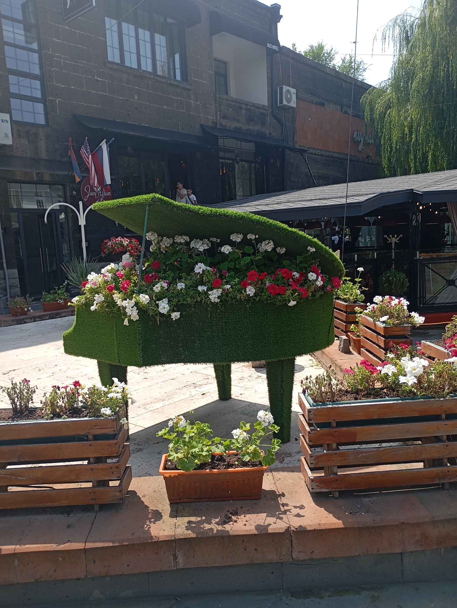 The image depicts an outdoor setting in Gyumri, Armenia, featuring a decorative piano sculpture covered in artificial grass, adorned with numerous blooming flowers. The piano sculpture is positioned in the foreground, with its lid open, showcasing a vibrant display of red and white flowers. Several wooden planters filled with greenery and flowers are placed around the piano sculpture and on a raised brick ledge.
In the background, a two-story building with dark stone cladding is visible. The building has several windows and an awning or roof extending over a section of the ground floor. Flags, including the American and Russian flags, are displayed near the building's entrance. A sign with text, partially obscured, is also visible. The overall environment suggests a public space, possibly a cafe or a street in a commercial area, given the presence of outdoor seating structures and architectural elements. The lighting indicates it is daytime, with shadows cast by the objects and trees.