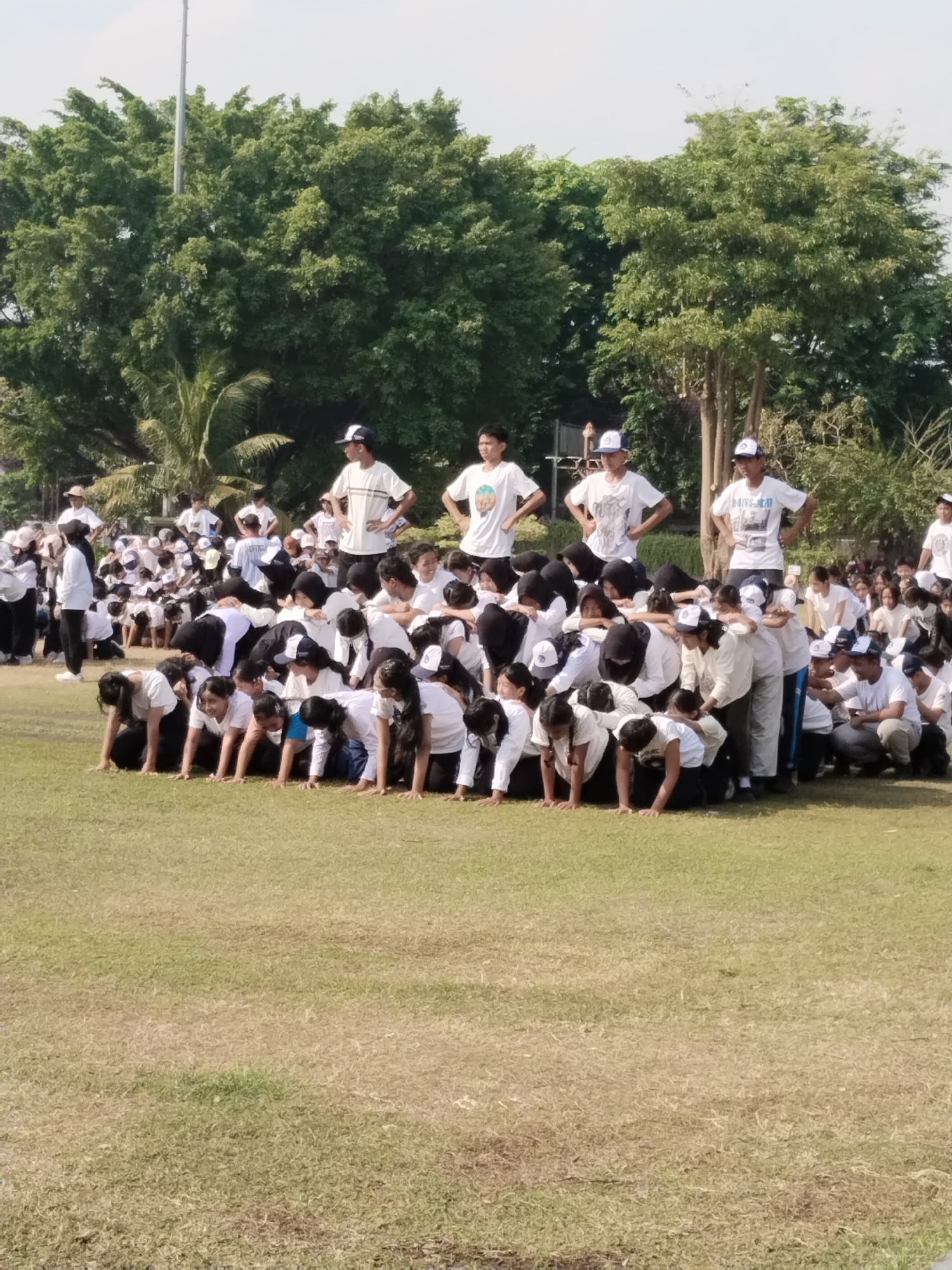 A group of approximately 50 to 70 people, predominantly young students in white t-shirts and black pants or skirts, are gathered on a grassy field in Kecamatan Pati, Indonesia. The majority of the individuals are in a kneeling or crouching position, forming a large, interconnected structure. Many are wearing white baseball caps. Some individuals are standing upright, observing the group. The environment is an outdoor setting with a clear sky and lush green trees in the background. The focus of the image is the human formation on the grass.