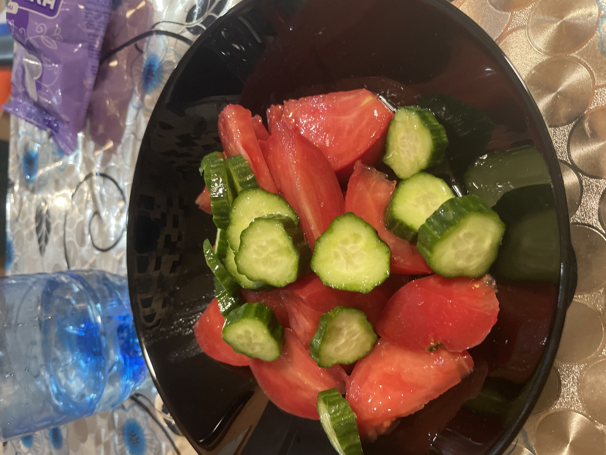A close-up image captures a fresh salad of sliced tomatoes and cucumbers served in a black bowl. The tomatoes are cut into large, vibrant red wedges, while the cucumbers are uniformly sliced into bright green rounds. Both vegetables appear glistening, suggesting freshness or the presence of a light dressing.
The bowl rests on a table covered with a tablecloth featuring distinct patterns. To the right of the bowl, the tablecloth displays a design of concentric circles, possibly metallic or reflective, in a golden or light brown hue. To the left, a different section of the tablecloth is visible, characterized by a white background adorned with blue floral patterns.
On the left side of the frame, a purple plastic bag is partially visible. The letters "HU" in white are discernible on the top left corner of the bag, with other unreadable text below it. The bag's contents appear to be purple items. Adjacent to the bag, a clear plastic water bottle, mostly empty, lies on its side, revealing blue reflections or elements within its structure.
The scene suggests a casual indoor setting, possibly a kitchen or dining area in Ihtiman, Bulgaria, where a simple meal or snack has been prepared. No individuals are present, and the time of day or weather cannot be determined from the photograph.