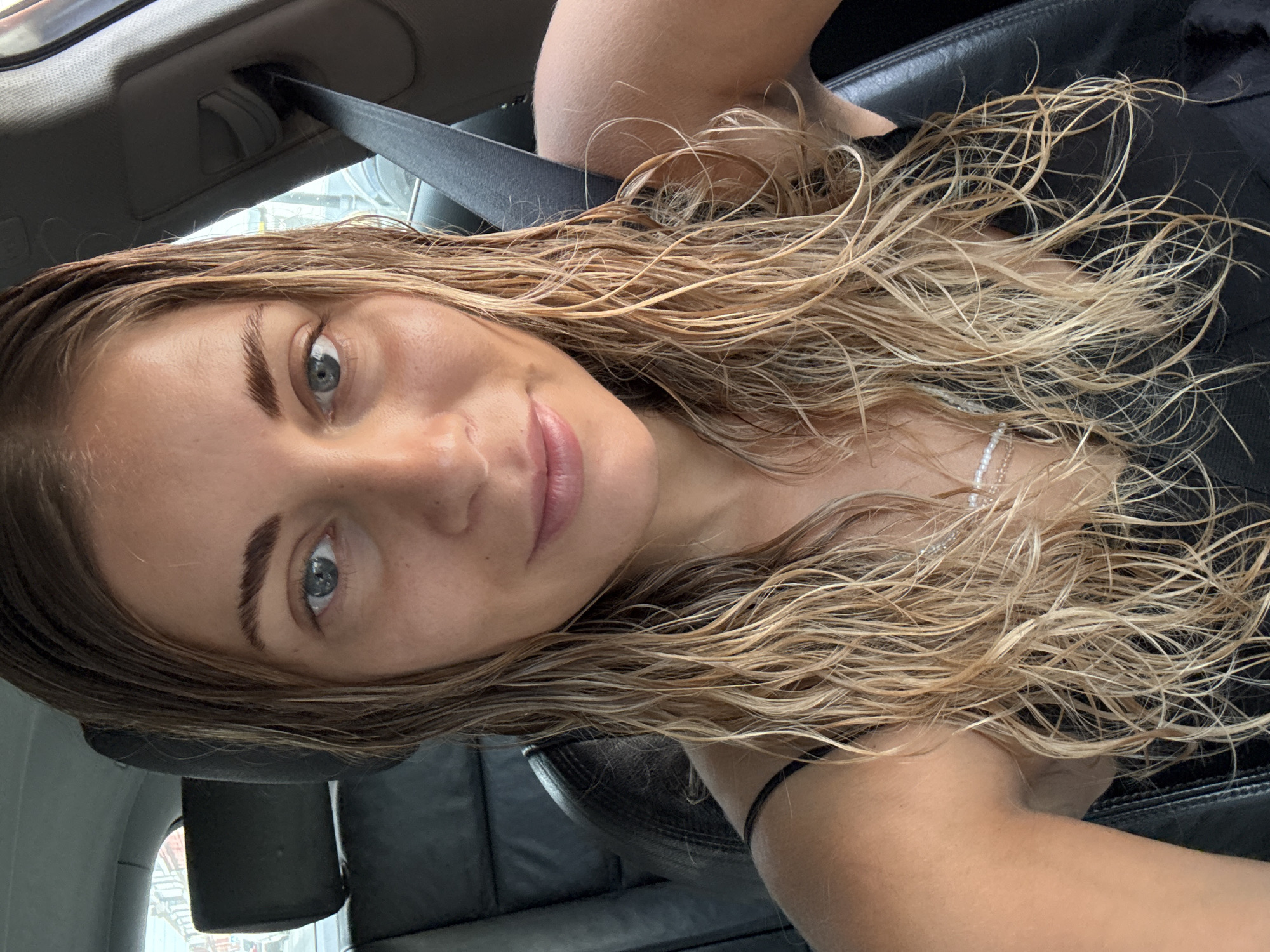 A young woman with wet blonde hair is seated in the driver's seat of a car, taking a selfie. She is wearing a black tank top and a delicate pearl necklace. The car's interior, including a black leather seat and a seatbelt, is visible. The background shows a blurred view of buildings and streetlights, suggesting an urban environment. The lighting indicates it might be daytime, possibly overcast or during the golden hour, given the warm tones on her skin. The overall impression is a candid moment captured within a vehicle.