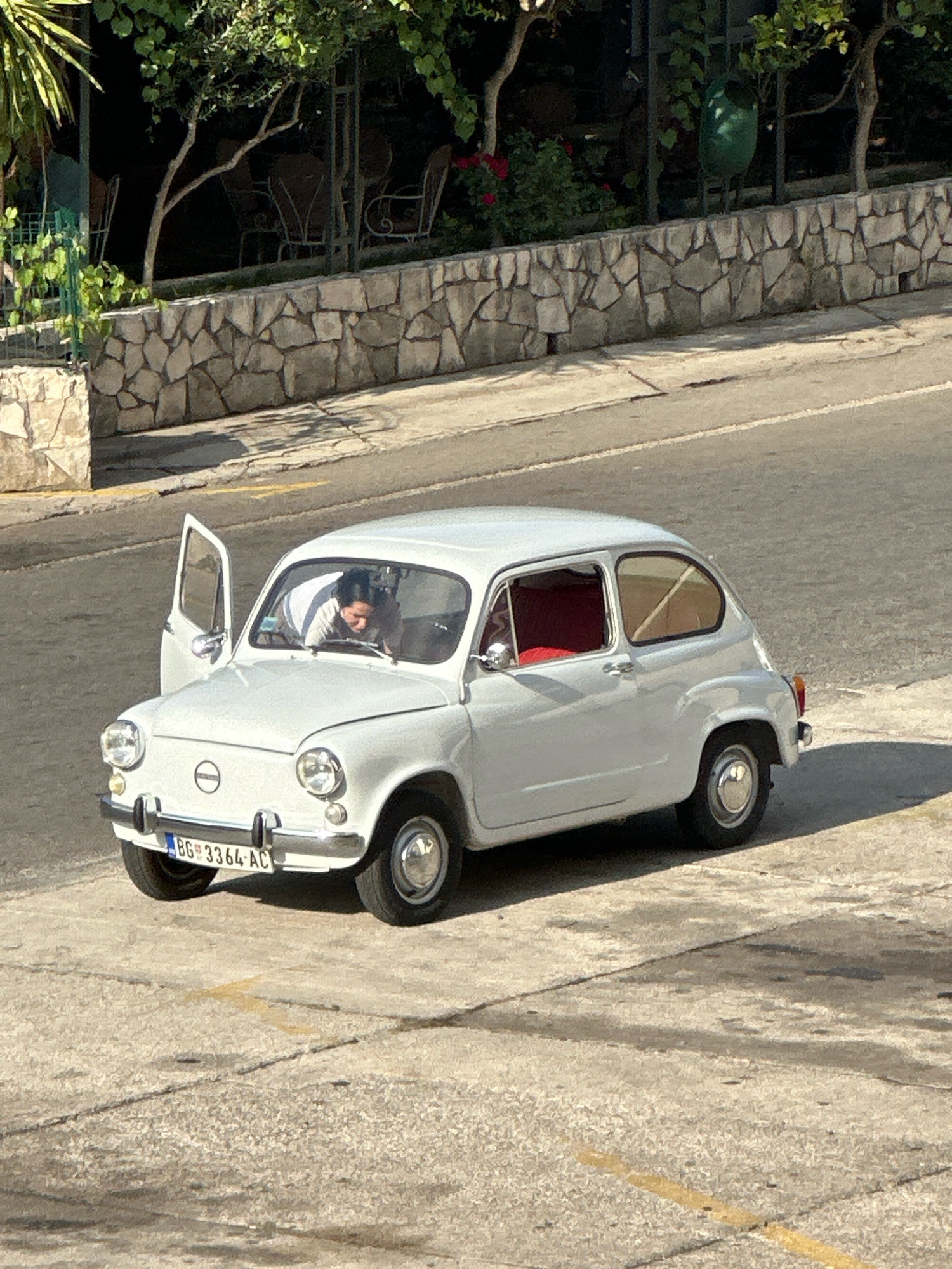 A white, vintage Fiat 600 car is parked on a concrete surface in what appears to be a residential or commercial area, possibly in Budva, Montenegro. The car's driver's side door is open, and a person is leaning forward from the driver's seat, appearing to be looking at something on the dashboard or floor. The car has a license plate that reads "BG 3364-AC". Behind the car, a stone wall is visible, with some greenery and trees above it, suggesting a hillside or elevated area. The lighting indicates it is daytime, with shadows cast by the car and surrounding objects. The environment appears to be an urban setting, with the car parked on a paved area that shows some cracks and discoloration.