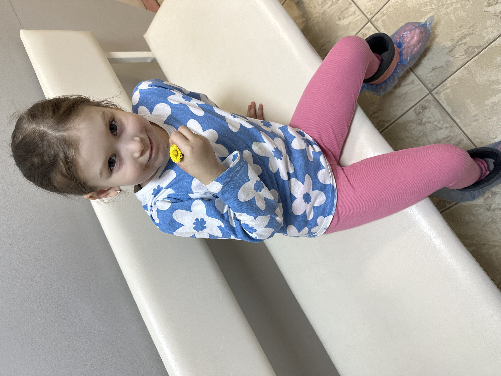A young girl, likely a toddler or preschooler, is seated on a light-colored, two-sectioned bench, looking directly at the camera with a gentle smile. She has light brown hair and dark eyes. She is wearing a blue long-sleeved shirt with a white floral pattern and pink leggings. Her feet are covered in dark shoes, which are further encased in clear plastic shoe covers. In her right hand, she holds a small yellow flower, possibly a dandelion.
The setting appears to be an indoor environment, with a plain light gray wall in the background to the left and a tiled floor with light-colored square tiles to the right. The bench itself is smooth and reflective, suggesting a material like vinyl, commonly found in waiting rooms or examination areas. The presence of shoe covers indicates a requirement for cleanliness, reinforcing the likelihood of a medical facility or similar public health setting, consistent with common practices in places like Chaikovsky, Russia. The girl appears calm and content. No text is visible in the image.
