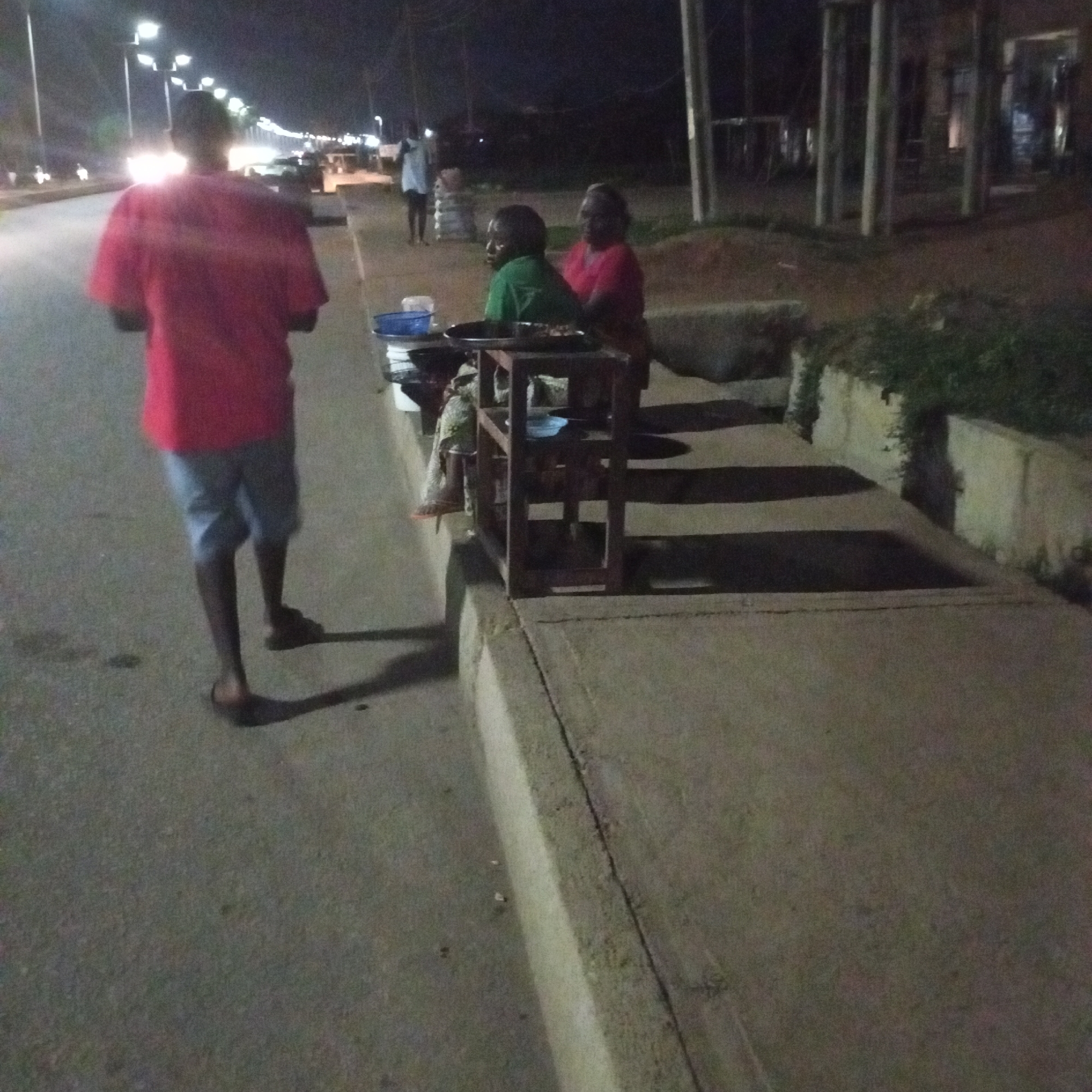 At night in Unknown, Unknown, a street scene shows a man in a red shirt and light blue shorts walking away from the camera on an empty roadway. A food stall is set up on the sidewalk, manned by two people, one in green and one in red. Streetlights illuminate the road, and there is visible vehicle traffic in the background. The scene includes buildings and greenery.