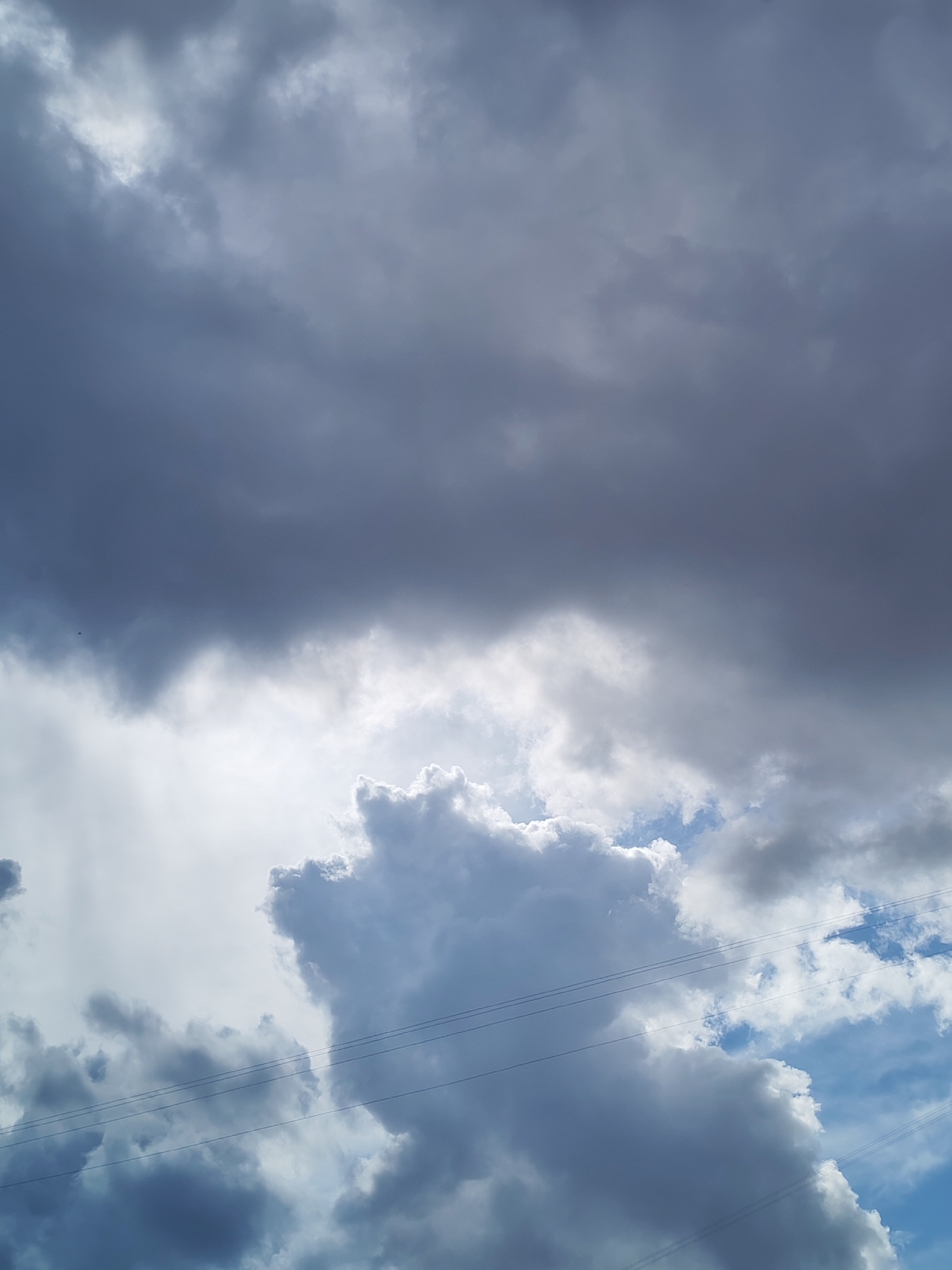 The image presents a vast sky over Izhevsk, Russia, showcasing a dramatic interplay of clouds and light. The upper and left portions of the frame are densely filled with thick, dark grey clouds, hinting at overcast conditions or impending rain. Toward the center and lower half of the image, the sky brightens significantly as a powerful light source, presumably the sun, breaks through the cloud cover. This light illuminates a large, prominent cumulus-like cloud with brilliant white edges and shaded blue-grey undersides, creating a striking contrast against the darker clouds. Patches of clear blue sky are visible in the lower right corner, suggesting a break in the weather or a partly cloudy day.
Multiple thin, dark power lines stretch horizontally across the lower middle section of the image, partially disappearing behind clouds and across areas of blue sky. These lines suggest an urban or suburban environment. The scene is captured during daylight hours, as indicated by the intense illumination. There are no people, other specific objects beyond the clouds and power lines, or any visible text in the photograph. The image conveys a sense of natural grandeur and the dynamic movement of atmospheric conditions.