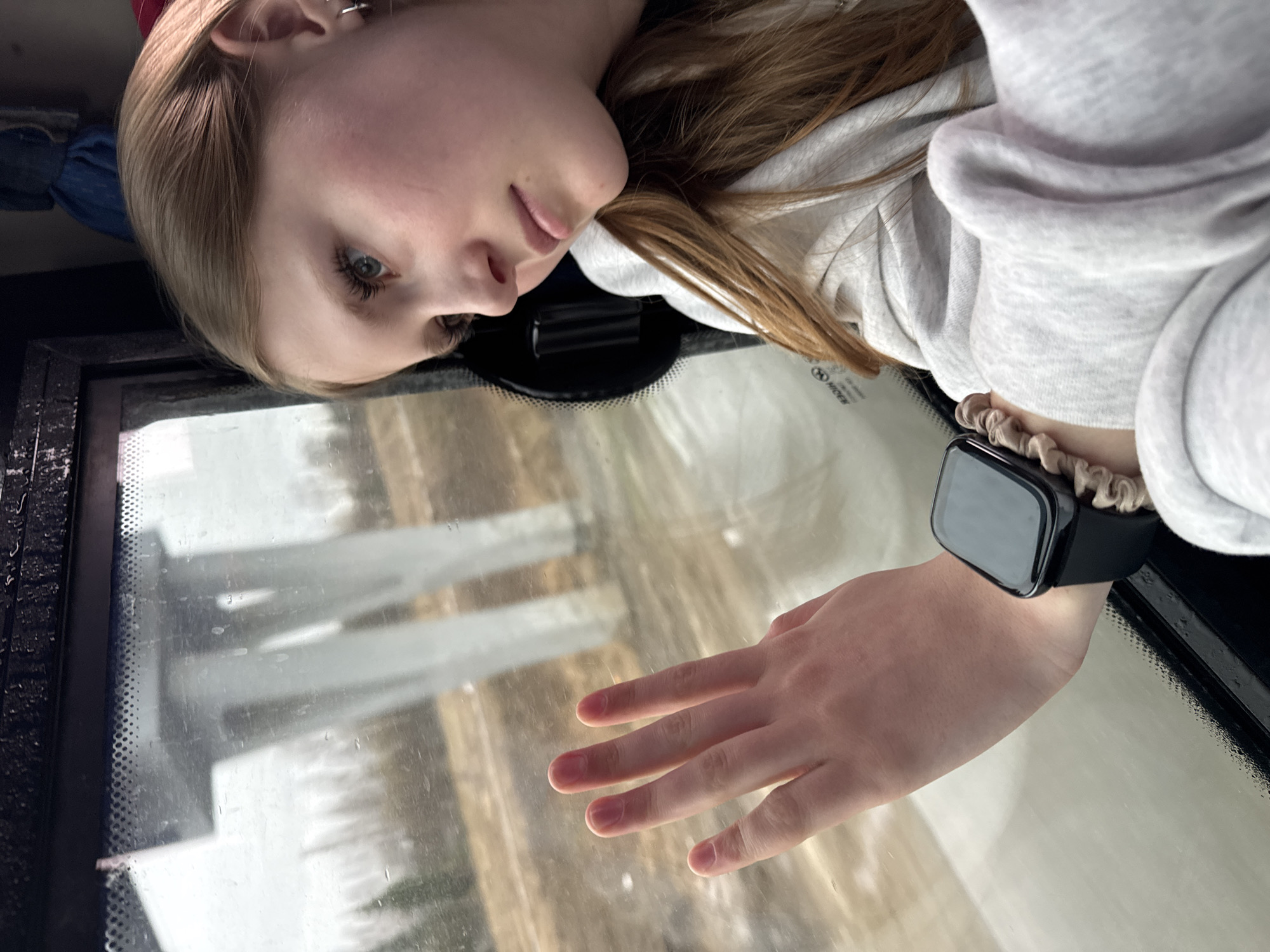 A blonde-haired woman with light skin is looking down in a photograph taken inside a vehicle. She is wearing a light grey hooded sweatshirt and an Apple Watch with a beige scrunchie around the wrist. Her hand is extended and resting on the windshield of the vehicle, which has water droplets on the glass. The reflection of a snowy urban environment can be seen in the windshield. The photo was taken inside the vehicle in Kukushtan, Russia.