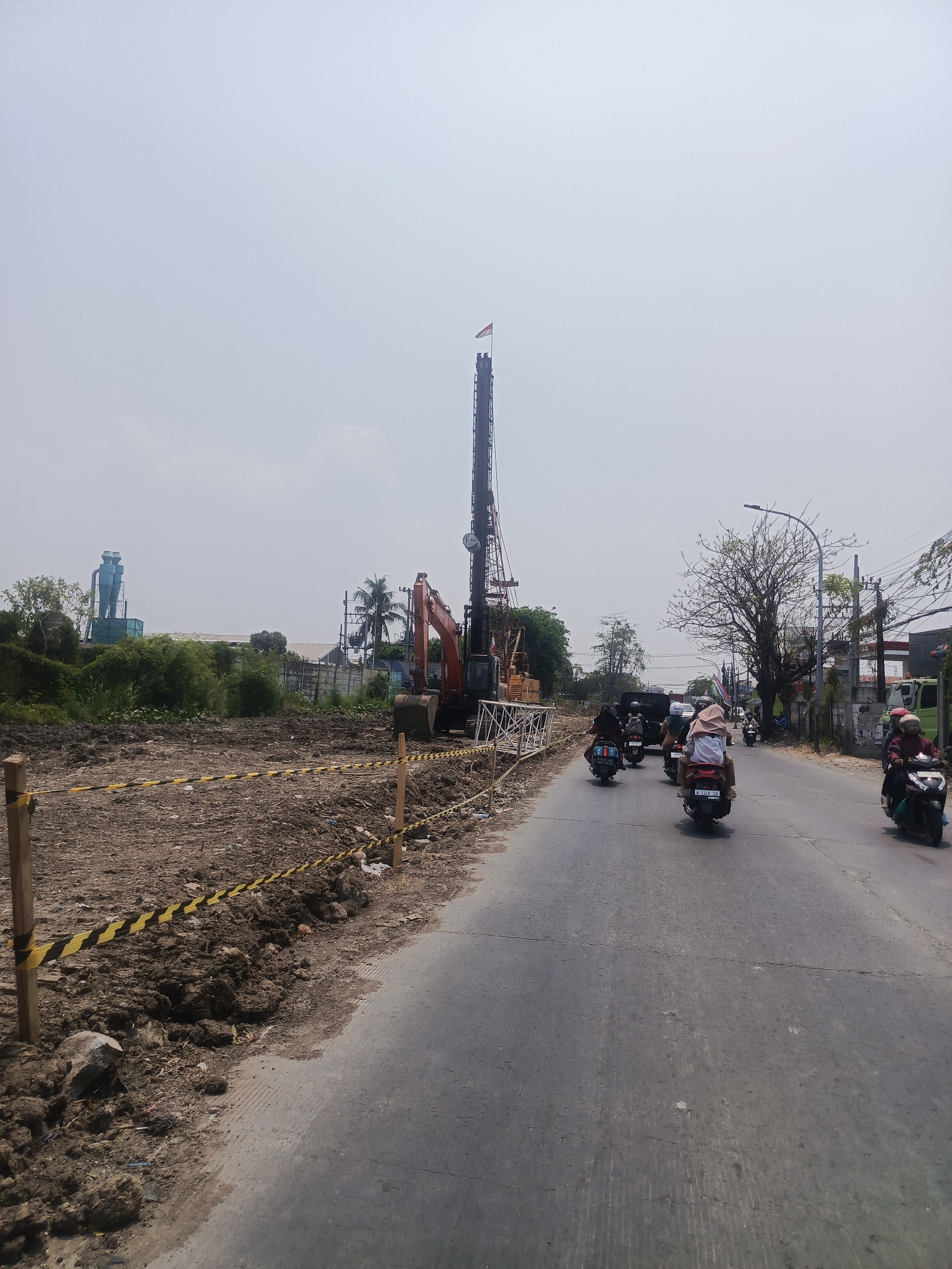 The image depicts a construction site on the side of a road in Surabaya, Indonesia. In the foreground, a dirt embankment marked with yellow and black caution tape lines the edge of the road. Several motorcycles with riders are traveling on the asphalt road, which appears to be a main thoroughfare. Further into the scene, a large construction rig with a tall mast stands on the dirt lot, with an excavator visible nearby. In the background, there are trees, buildings, and a blue industrial structure. The sky is overcast. The construction site suggests ongoing development or infrastructure work within the city.
