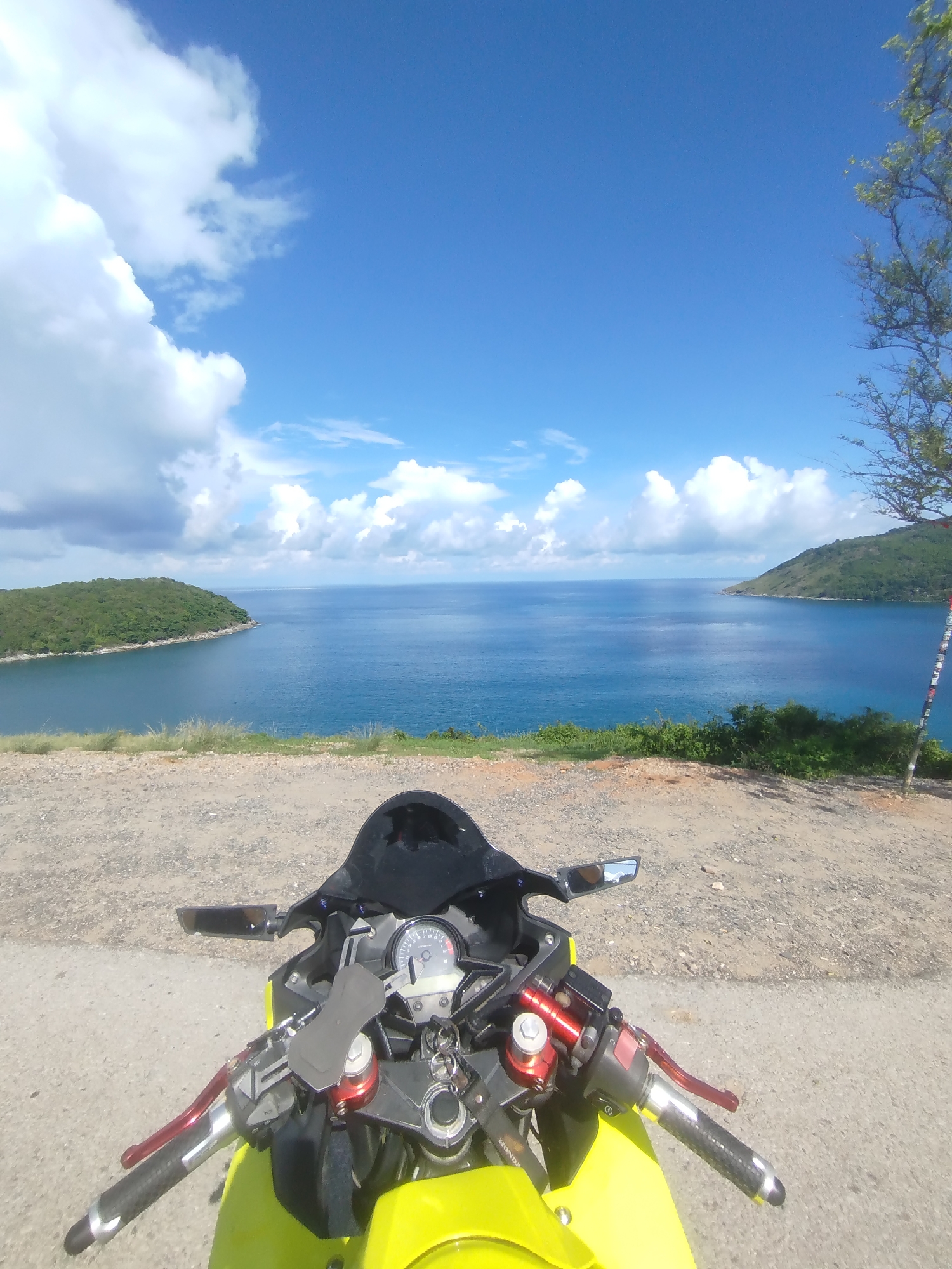 A first-person perspective shot shows the handlebars and front of a bright yellow motorcycle parked on a gravel road. The motorcycle's dashboard and black windscreen are visible. Beyond the motorcycle, a scenic view unfolds of a bright blue ocean meeting a similarly blue sky dotted with fluffy white clouds. Two lush green peninsulas jut out into the water on either side of a calm bay. The image appears to be taken in Rawai, Thailand, given the distinctive coastline and tropical vegetation.