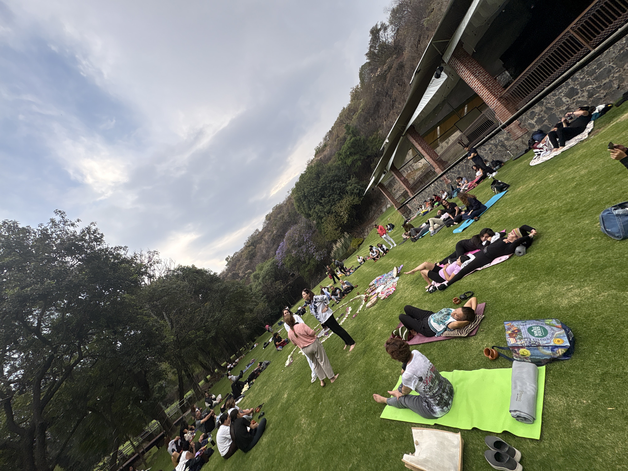A wide, canted shot depicts a group of people on a grassy slope, seemingly engaged in an outdoor activity. The location is identified as Santa Ana Acozautla, Mexico.
People are scattered across the lawn, some lying down on mats, others sitting, and a few standing. Two individuals are positioned in the center, potentially leading the group in an exercise or demonstration. Various mats, towels, bags, and personal items are spread around the area.
A building with a long, sloping roofline runs along the right edge of the frame, backed by a stone wall. Behind the building, a hillside covered with trees is visible. To the left, trees provide a natural backdrop. The sky is overcast with scattered clouds.