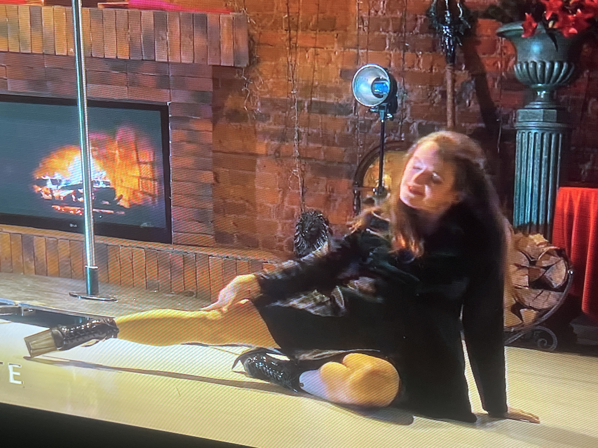 A woman is sitting on the floor in front of a brick wall. The wall is decorated with what appears to be fairy lights and some hanging vines. To the left of the woman is a television screen displaying a fireplace. A metal pole stands vertically next to the television. In the background, to the right of the woman, is a large urn containing red flowers and next to that, a pile of logs. The woman is wearing a black dress and black boots. She is looking down and to her right. The floor is a pale color with a textured pattern. This image does not provide any information about the city of Zoriane, Ukraine.