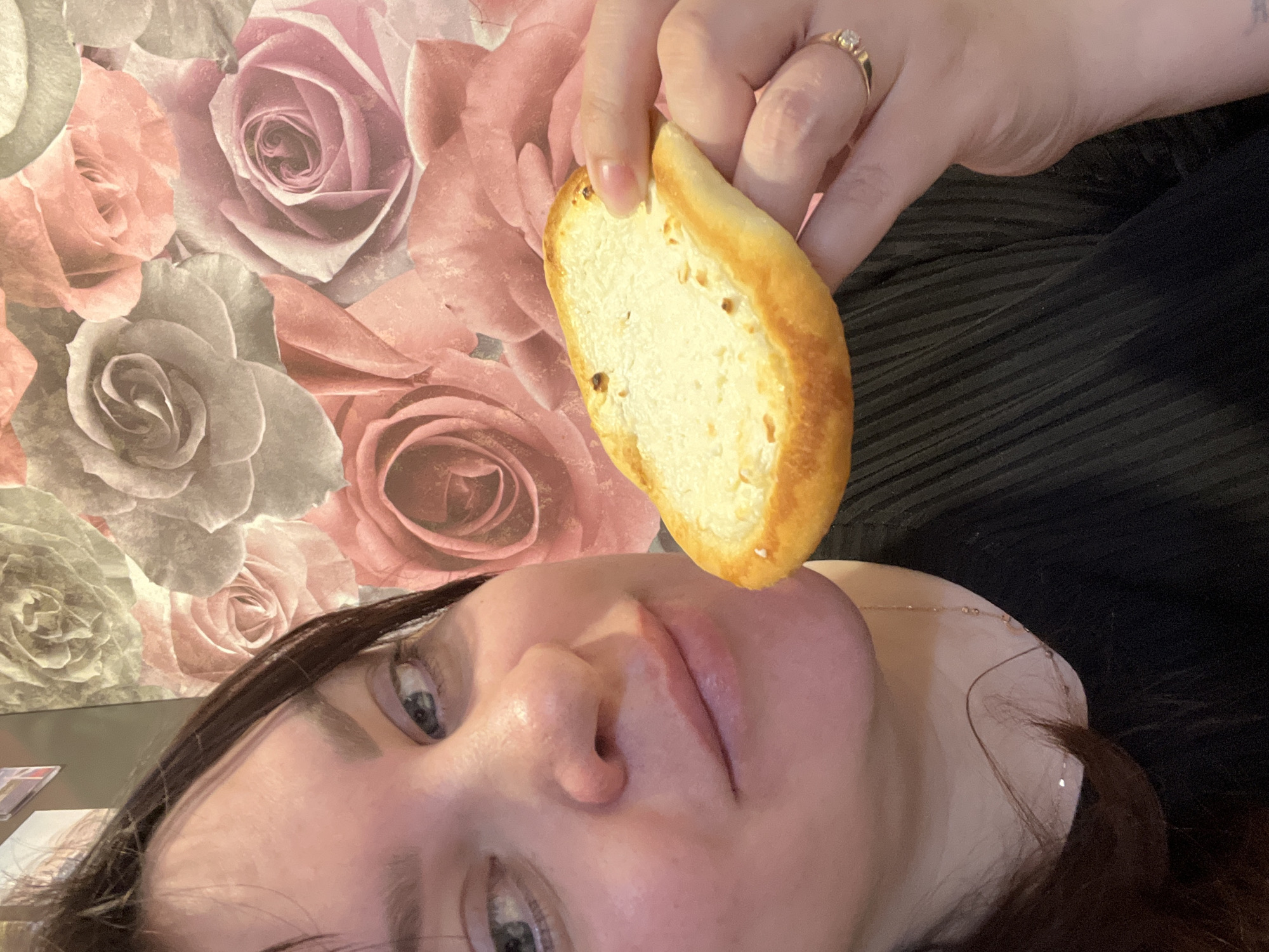 A woman with dark hair is holding a slice of bread in front of her face. She has light eyes and is wearing a gold necklace. She is wearing a ring on her finger. The background is a wallpaper with roses. The location is not mentioned in the image.