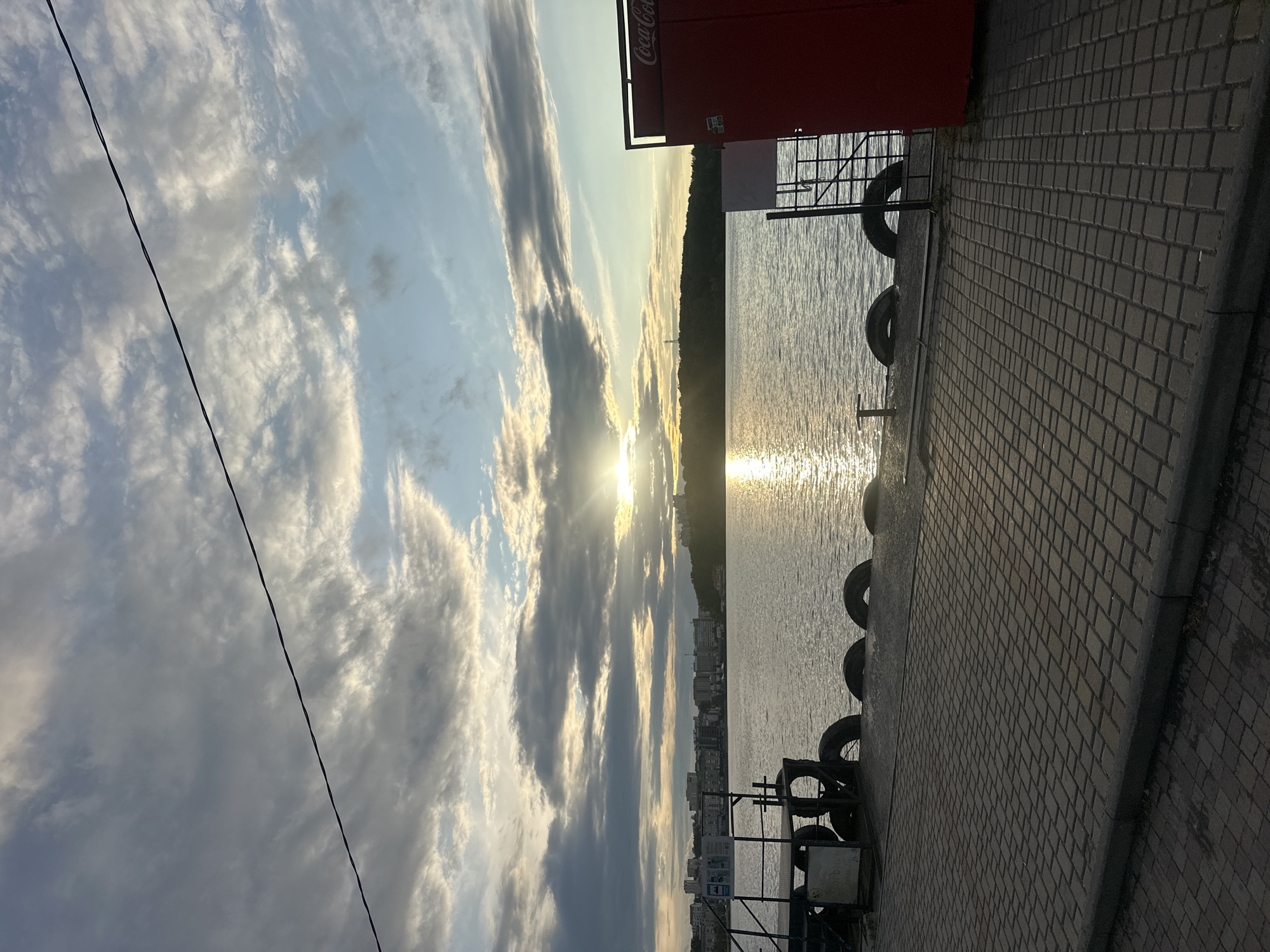 This is a photograph of a waterfront scene at sunset, likely in Ternopil, Ukraine. In the foreground, a paved walkway made of rectangular bricks leads towards the water. Along the edge of the walkway, several car tires are arranged, serving as a barrier or mooring. To the right, a bright red, rectangular structure with "Coca-Cola" branding is visible. The middle ground features a body of water, possibly the Ternopil Reservoir, reflecting the warm glow of the setting sun, which is partially obscured by clouds. In the distance, the cityscape of Ternopil is visible with buildings and structures lining the shore, and a hill covered in trees forms the background. The sky above is filled with a mix of dramatic clouds, with rays of sunlight breaking through, creating a visually striking sunset. There are no people visible in the image.