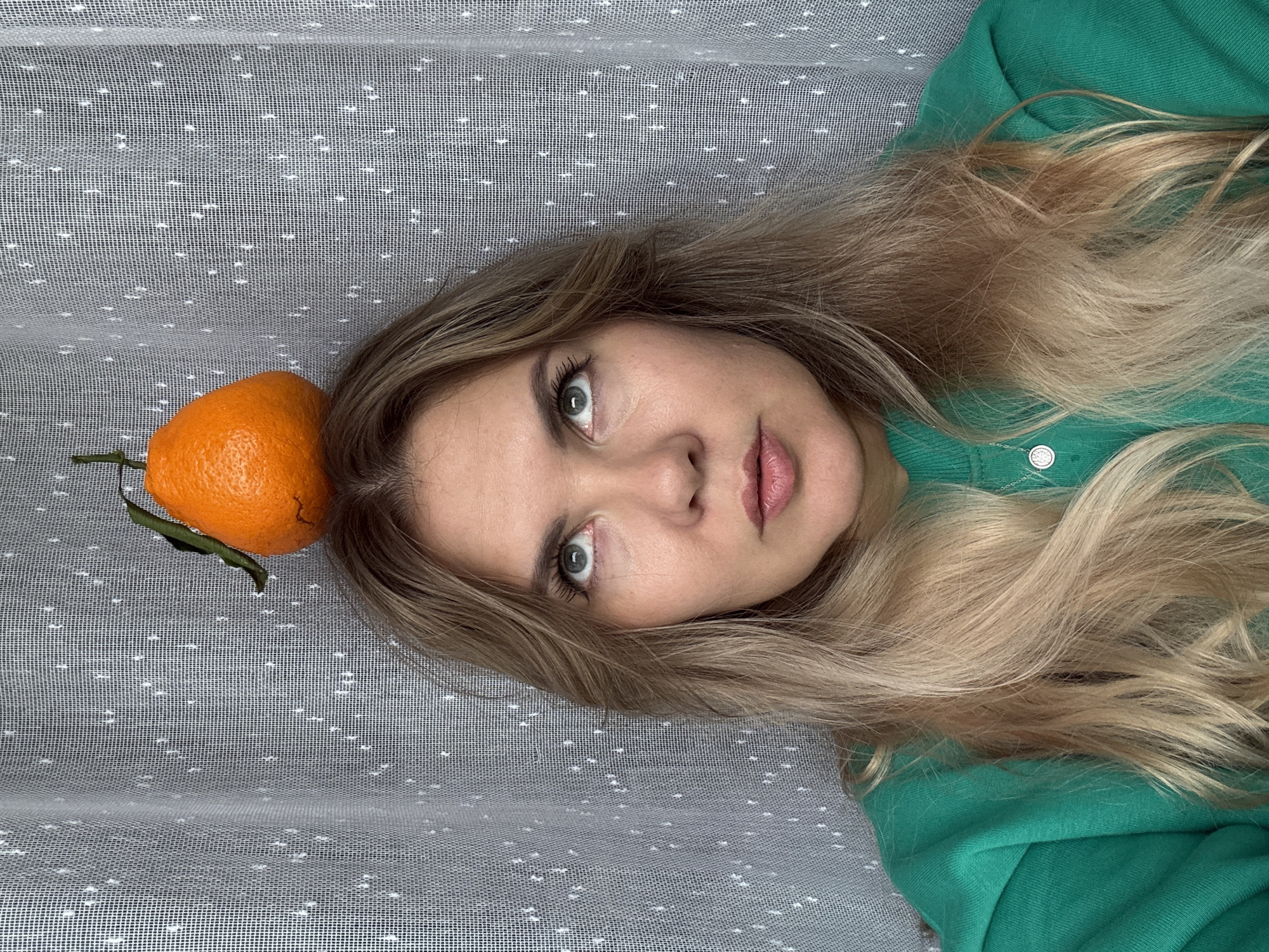 A medium shot shows a blonde woman with her eyes looking upwards, a bright orange tangerine balanced precariously on top of her head. She wears a green sweatshirt. The background consists of a white, semi-transparent curtain with a subtle dotted pattern. The lighting is soft and even. The setting appears to be indoors, likely a domestic environment. There is no discernible indication of a specific location within Izhevsk, Russia.